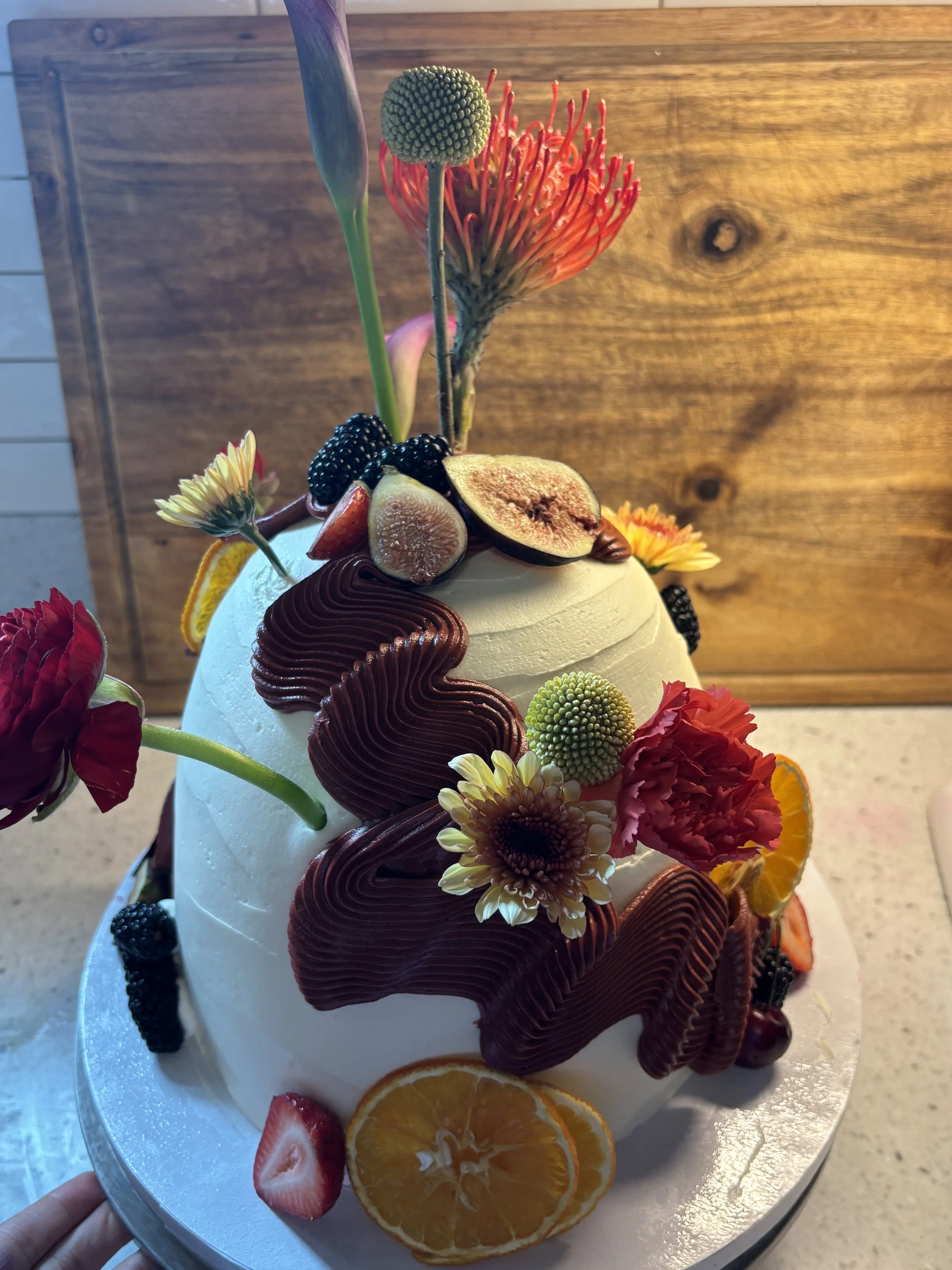 A cake decorated with fresh fruits, flowers, and frosting, with sliced oranges, strawberries, blackberries, and figs on top, on a glass tray against a wooden background.