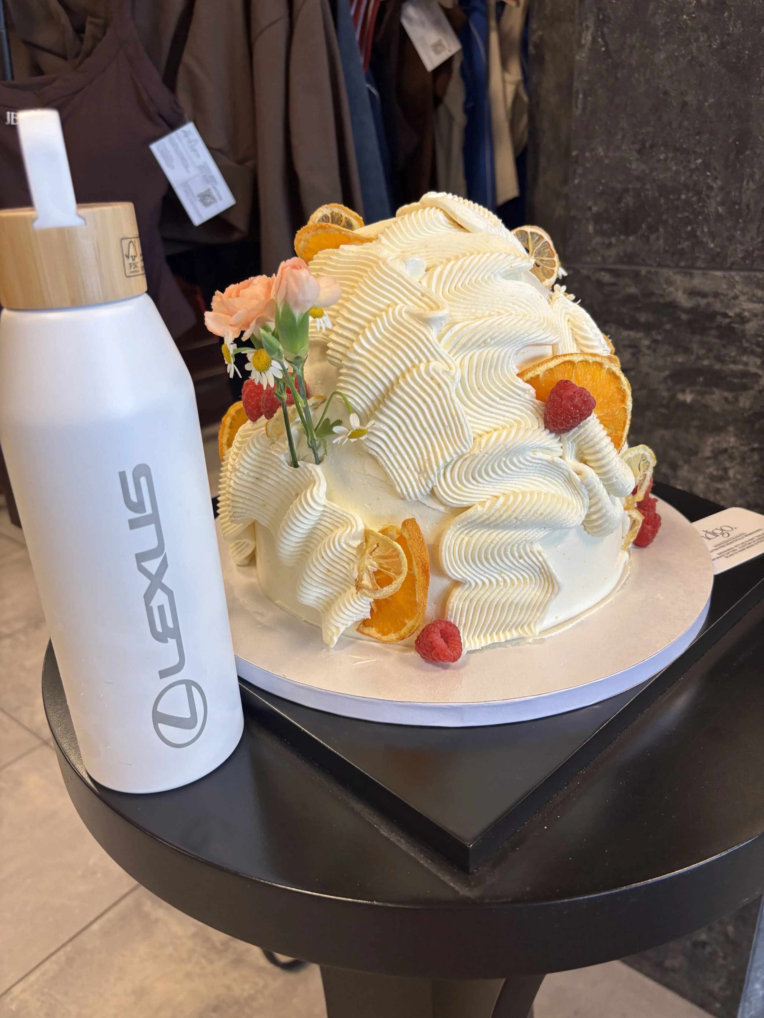 A decorative cake with white frosting, orange slices, raspberries, and flowers on top, placed on a black round table next to a white insulated bottle.