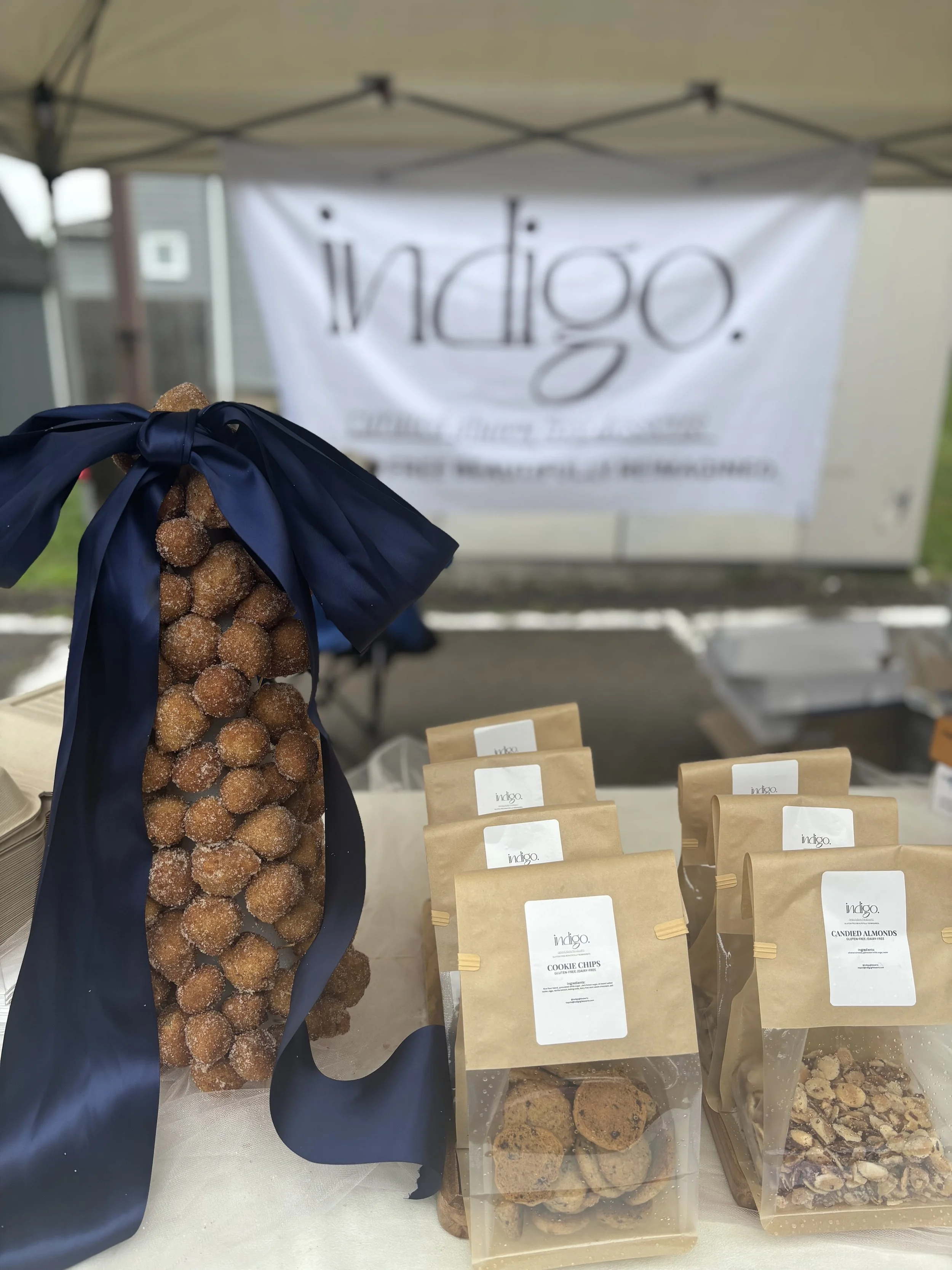 A display of various types of cookies and almonds in paper bags at an outdoor market stall, with a banner in the background reading 'indigo.'