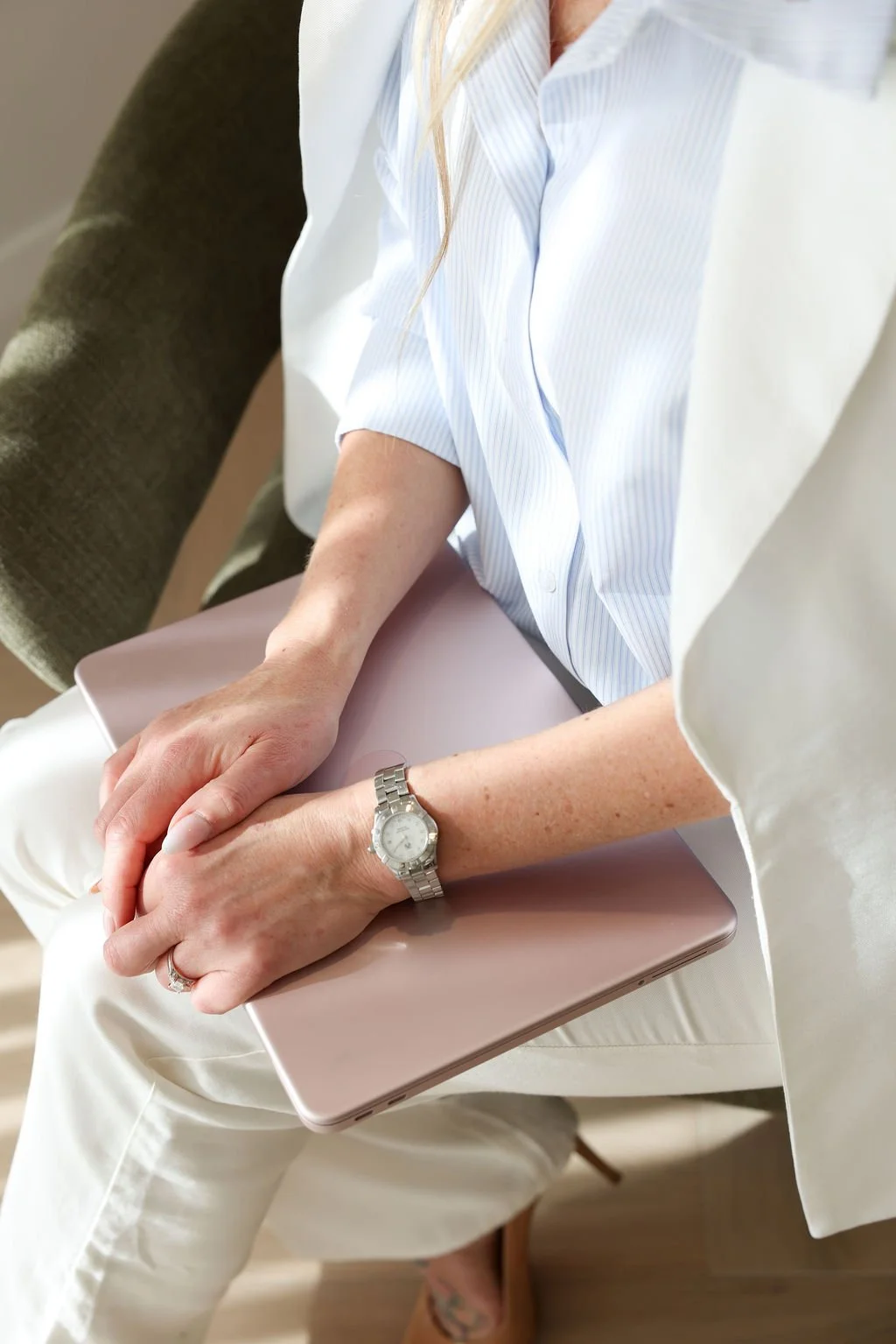 Close-up of a person's hands resting on their lap, holding a closed pink laptop, with a silver wristwatch on the left wrist, seated on a chair in a light-colored room.