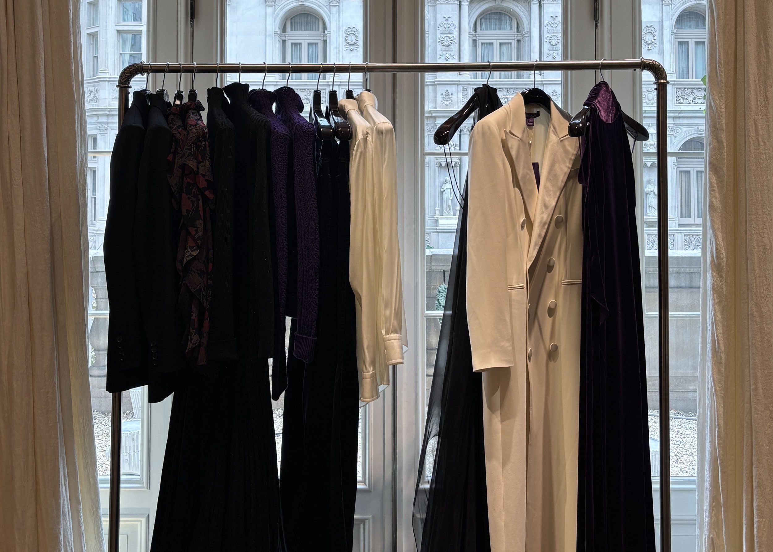 A clothing rack with various black, purple, and cream-colored dresses and coats in front of large windows with decorative architectural details.