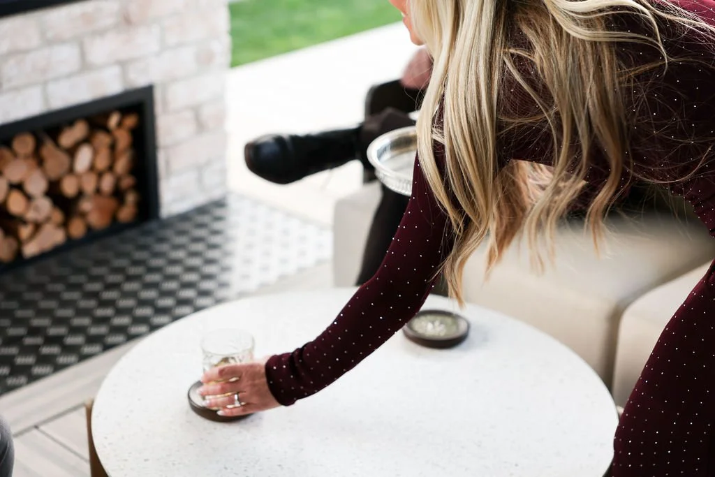 A woman with long blonde hair in a maroon dress with white polka dots is reaching for a glass on a white round table. There is a fireplace with a wood storage compartment to the left and a telescope in the background.