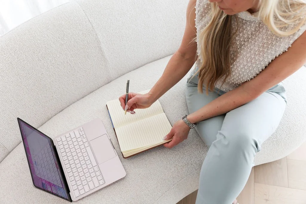 A woman sitting on a light-colored sofa, writing in a notebook with a pen, with a laptop open beside her.