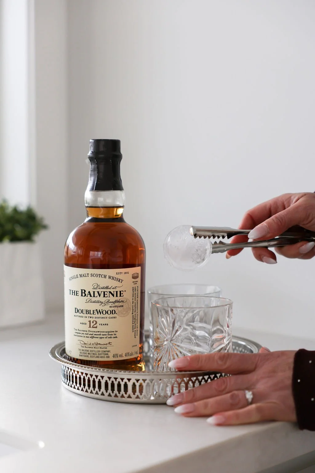Person using tongs to add ice to a glass with a bottle of The Balvenie Doublewood 12-year-old single malt Scotch whisky on a decorative tray.