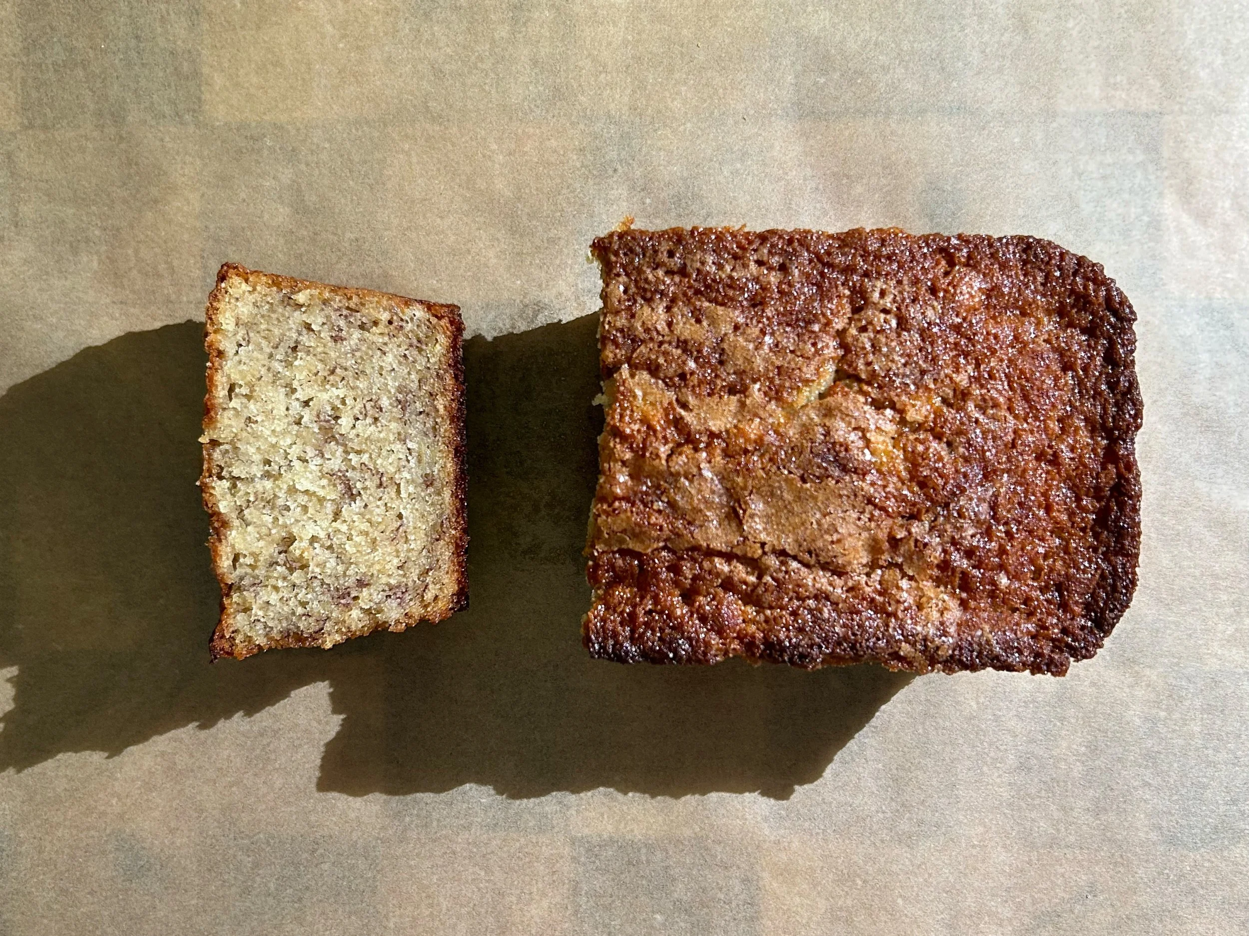 Close-up of two rectangular slices of banana bread, one lighter and one darker, placed on a beige surface with shadows underneath.
