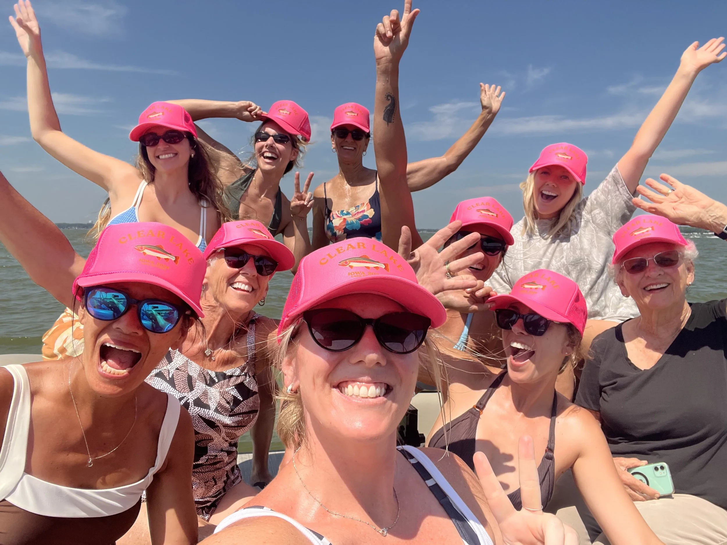 A group of smiling women wearing pink hats and sunglasses at the beach, enjoying a sunny day and taking a selfie together.