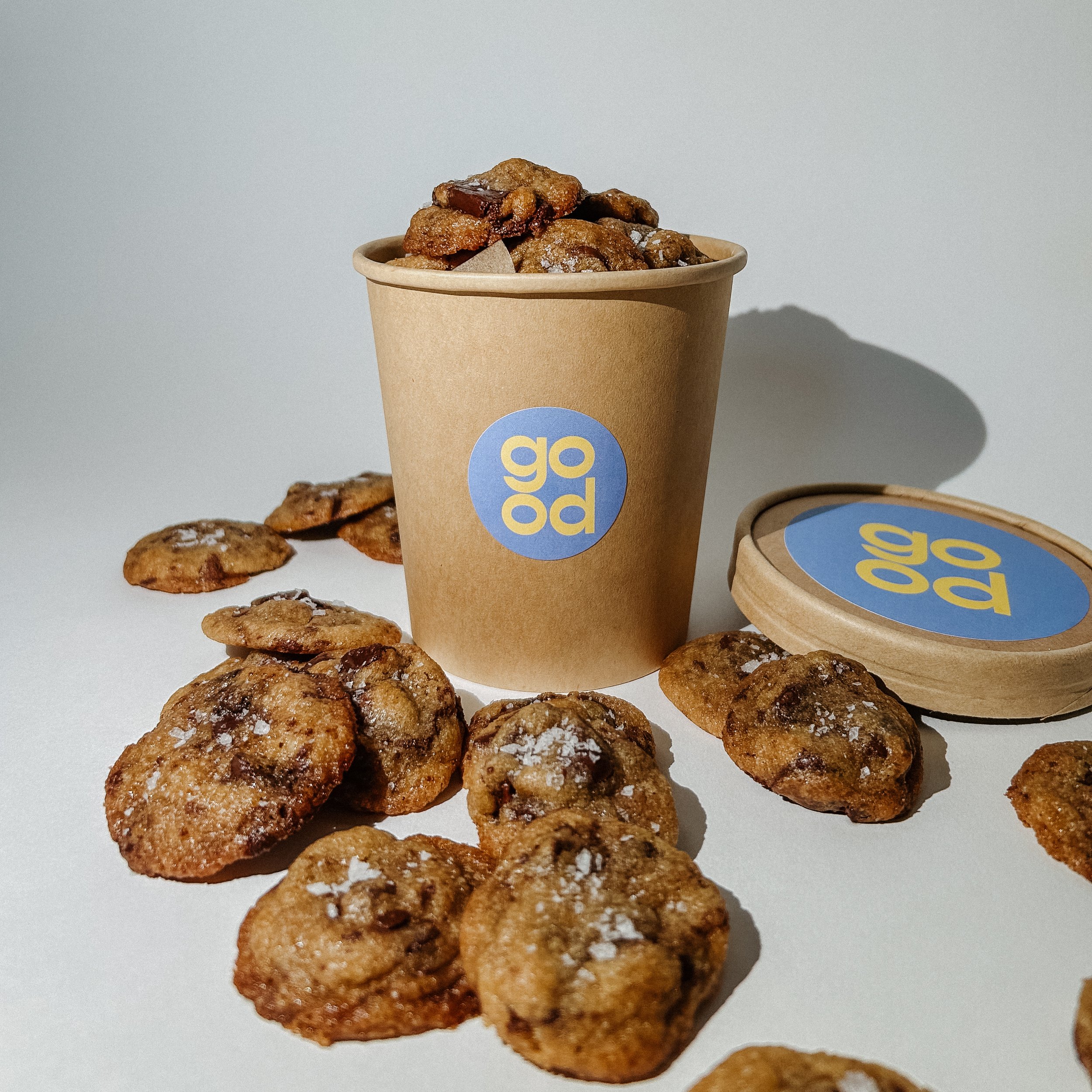Close-up of a brown paper cup filled with chocolate chip cookies and scattered cookies beside it on a white surface with a light gray background.