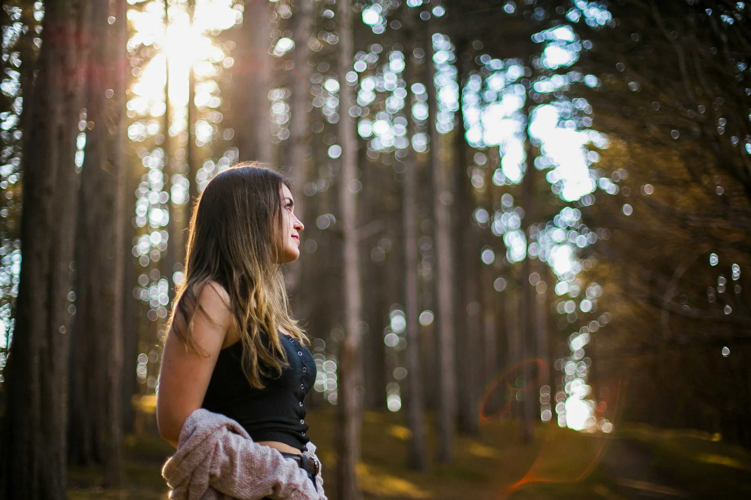 A woman with long brown hair standing outdoors in a forest, with sunlight filtering through trees in the background.