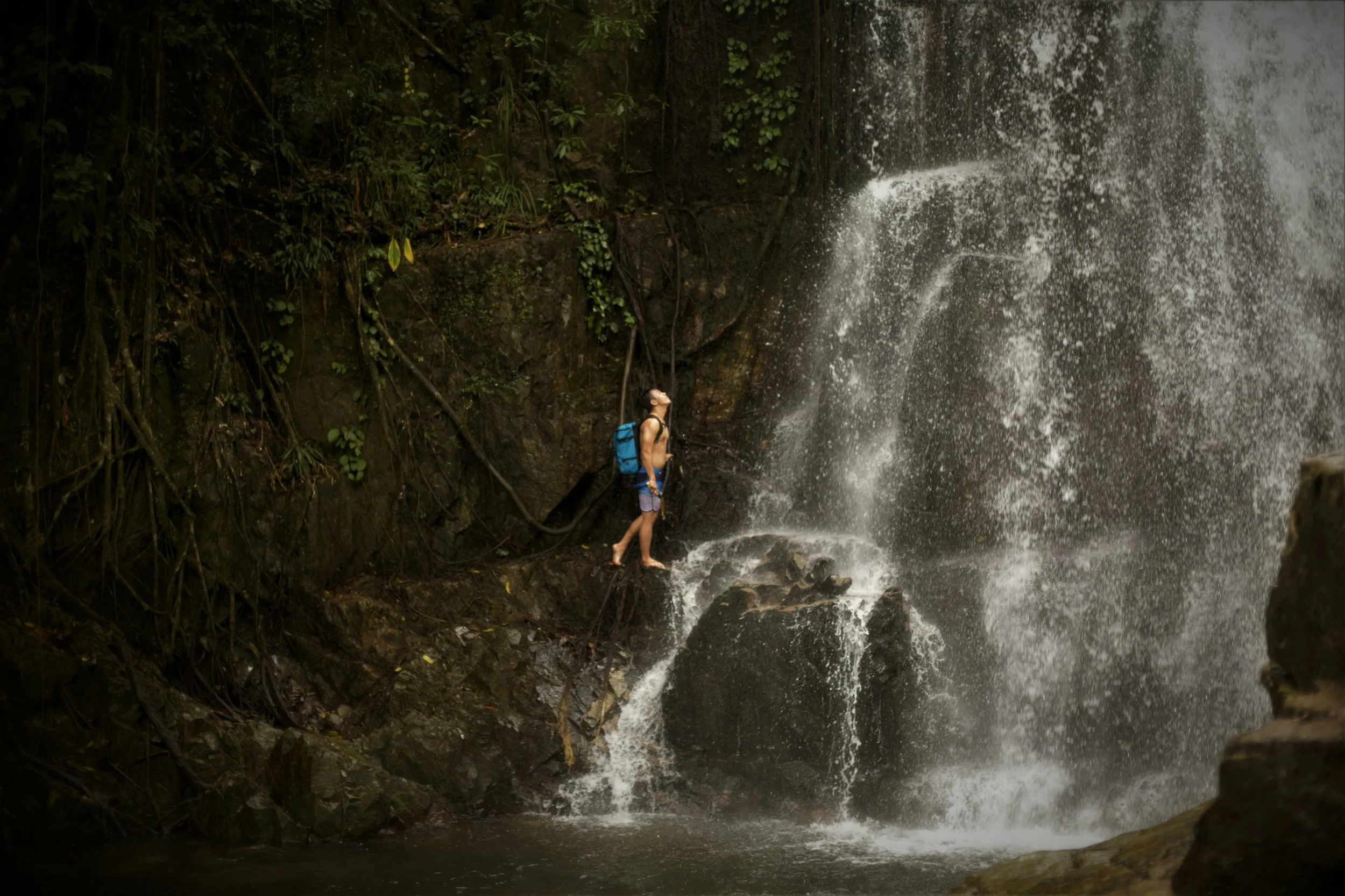 Person with a backpack standing on rocks in front of a waterfall in a jungle.