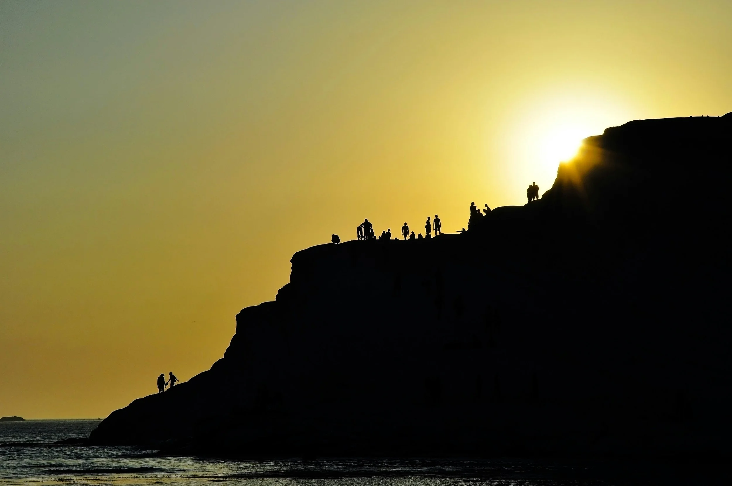 Silhouettes of people on a rocky cliff at sunset with the sun near the horizon.