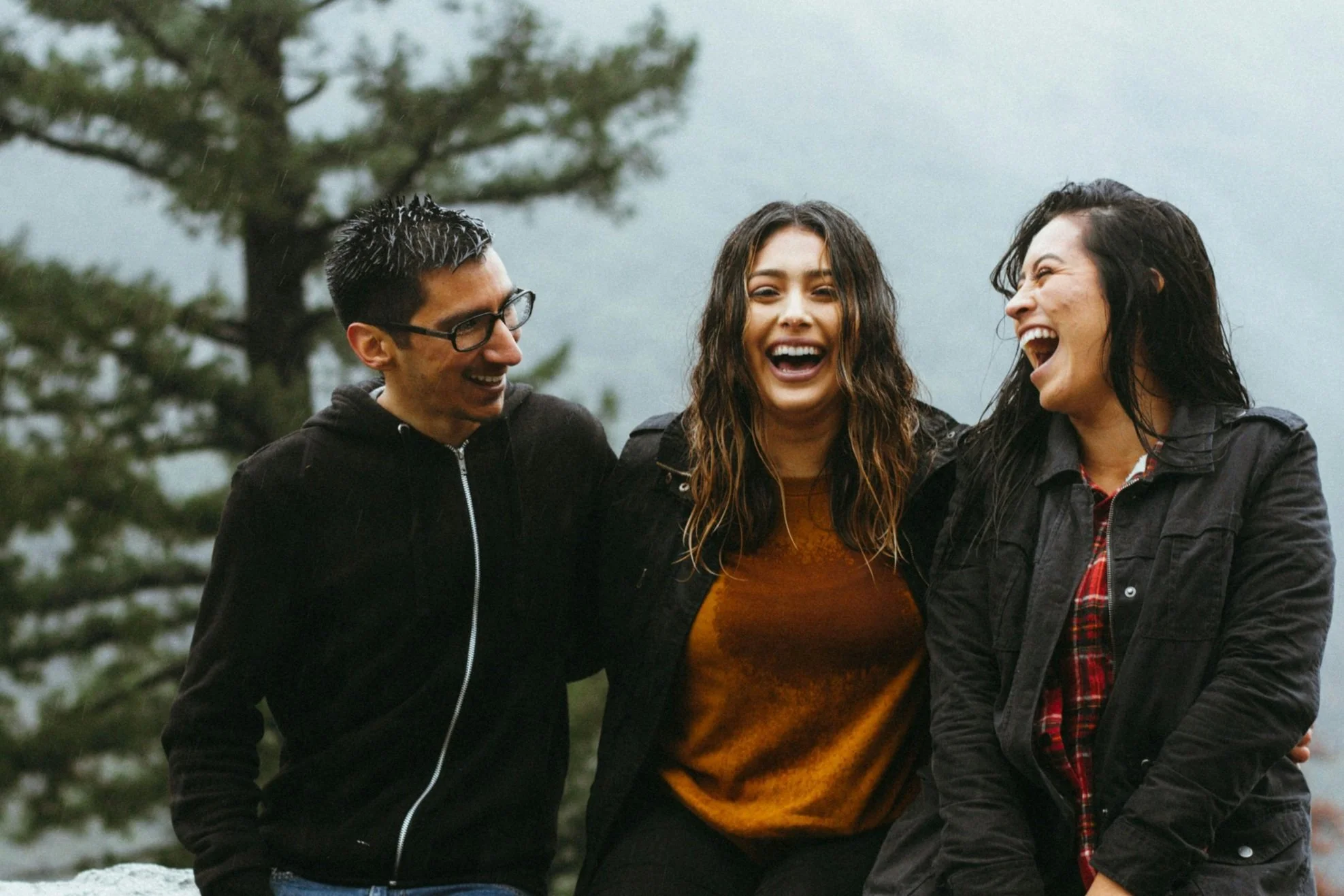 Three people outdoors, smiling and laughing, with trees and cloudy sky in the background.