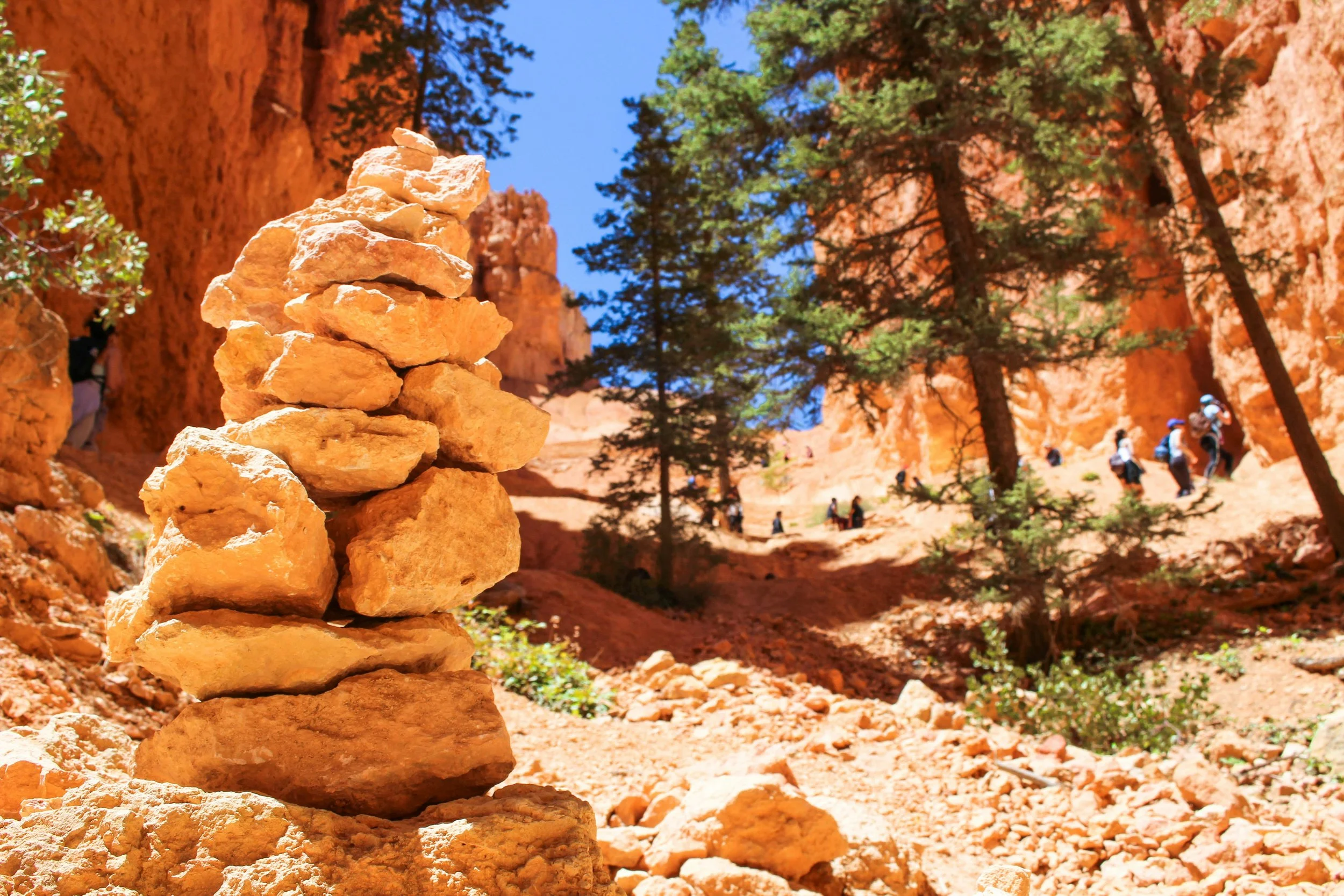 A stacked cairn of rocks in a desert canyon with trees and hikers in the background.