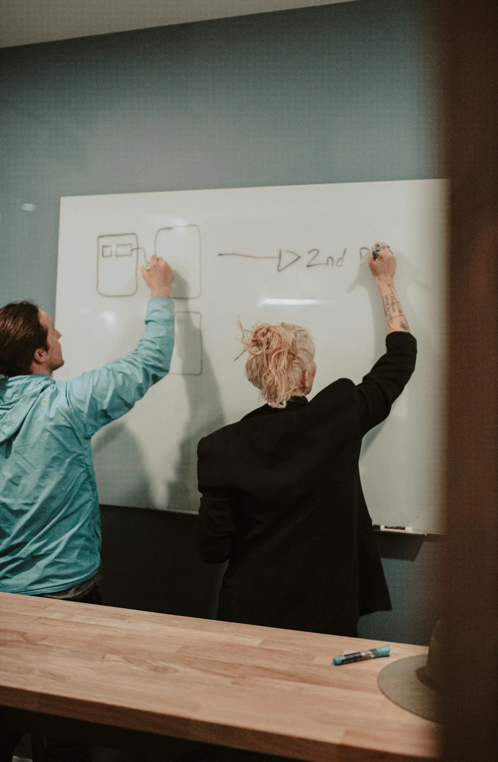 Two people, a man and a woman, are writing on a whiteboard; the man is drawing a computer, and the woman is writing text, with the word "second" visible.