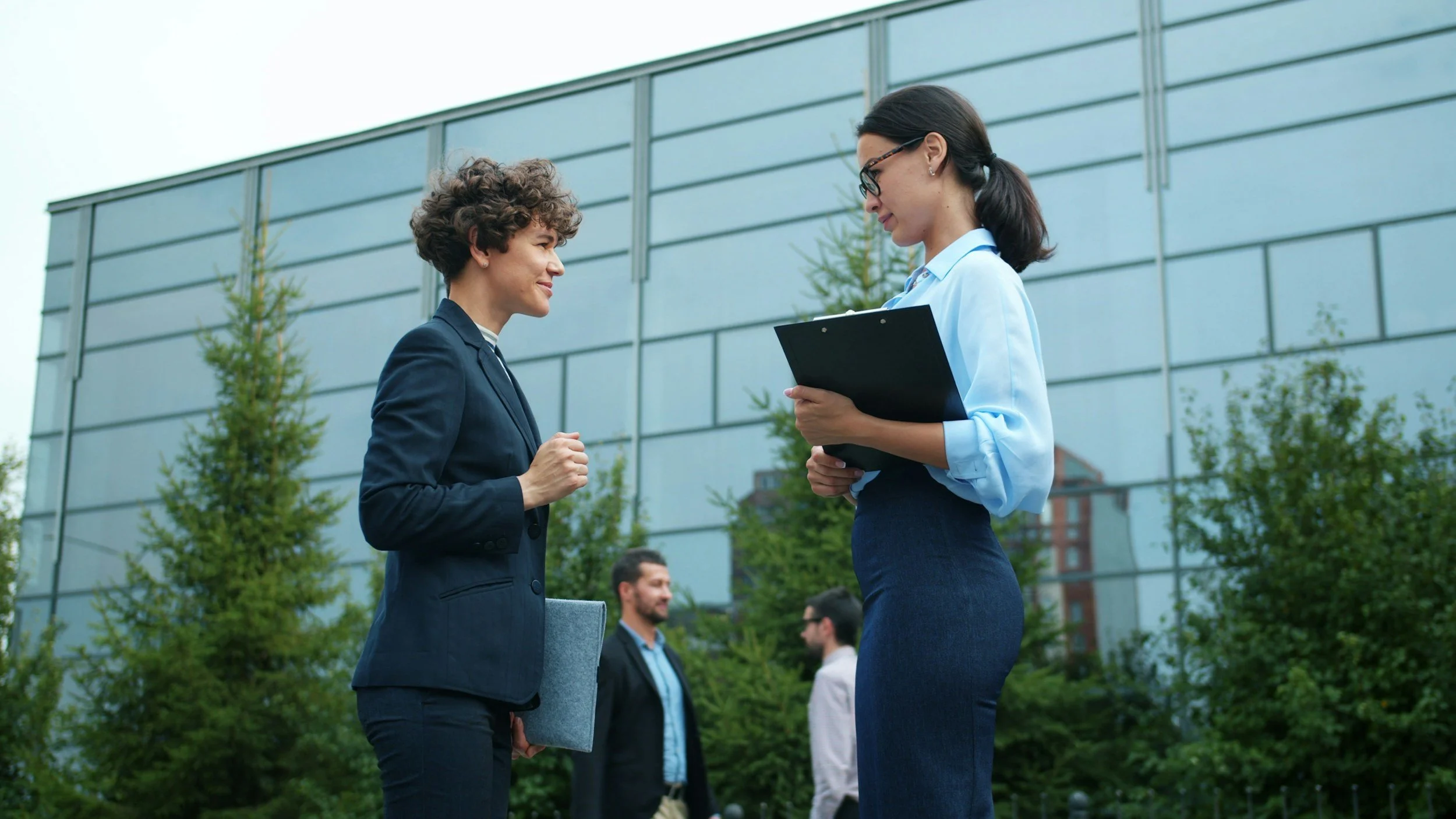Two women are having a conversation outdoors near a modern glass building. One woman is wearing a dark blazer and holding a folder, while the other woman, wearing glasses and a light blue shirt, is listening attentively.