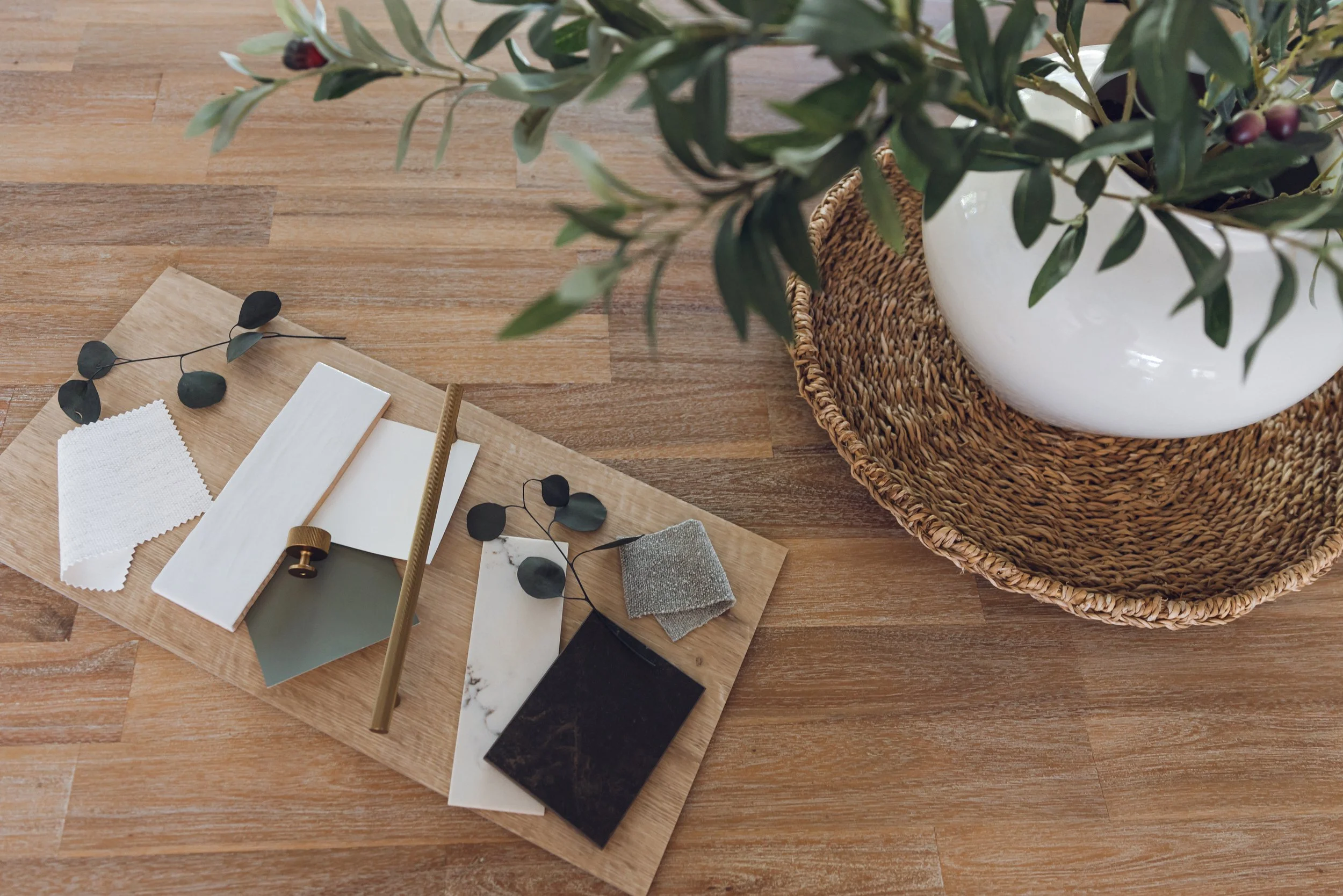 Flat lay of design materials on a wooden surface, including tiles, fabric samples, color swatches, and greenery in a white vase.