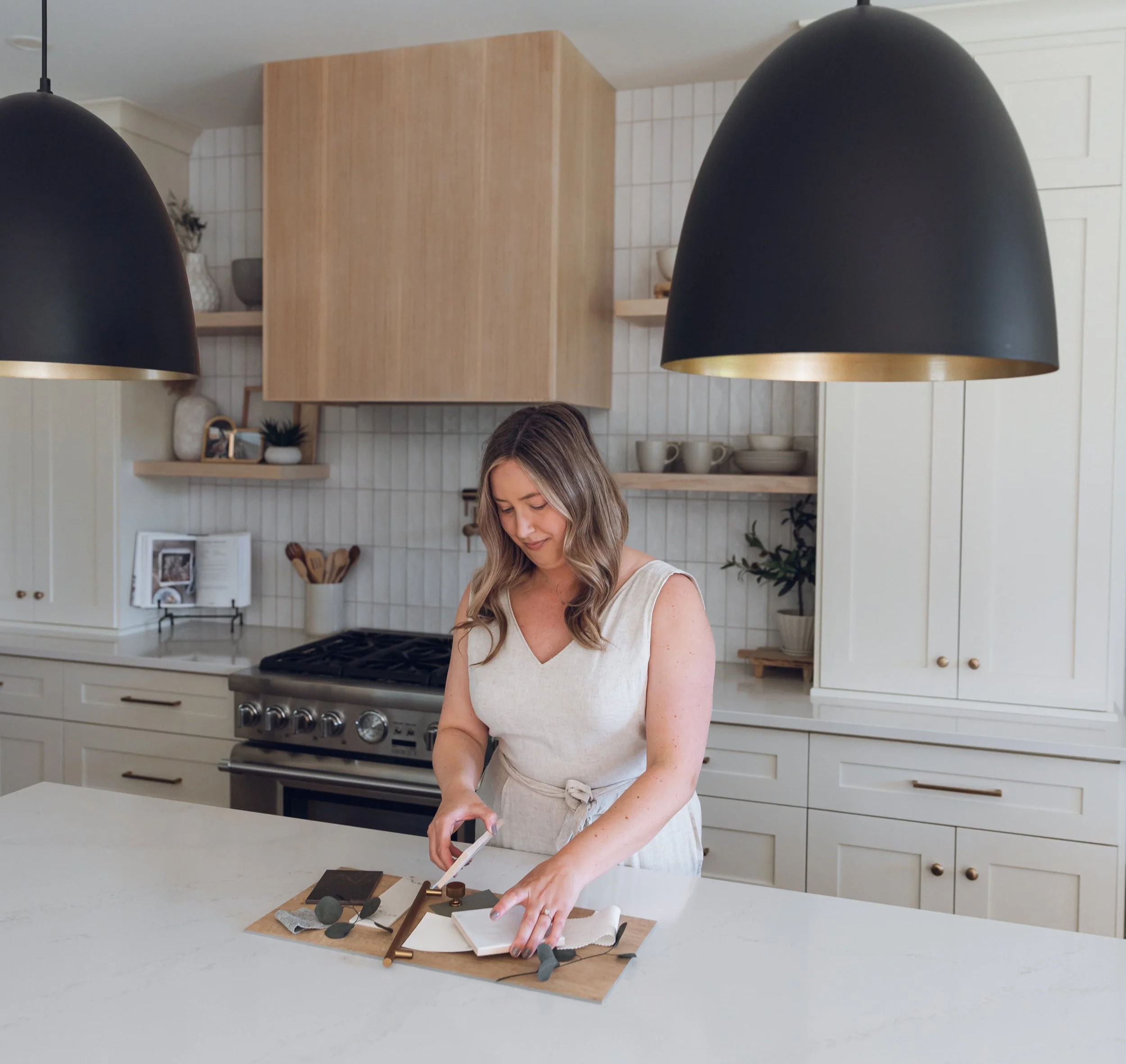 Alyssa in white dress preparing coffee in modern kitchen with hanging black pendant lights and wooden cabinets.