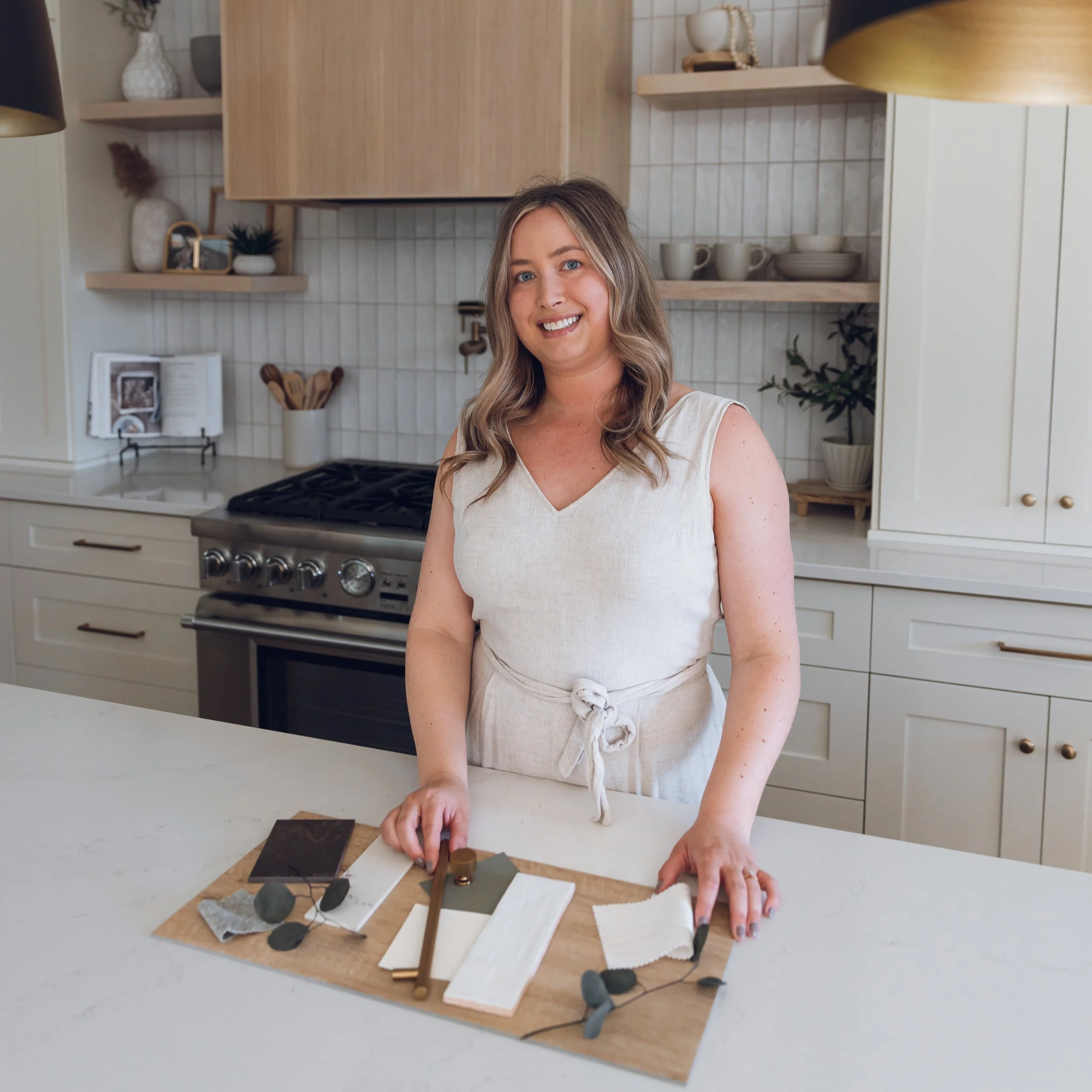 Alyssa in a cream dress smiling in a kitchen with open wooden shelves, a stove, and a countertop with fabric swatches and design samples.
