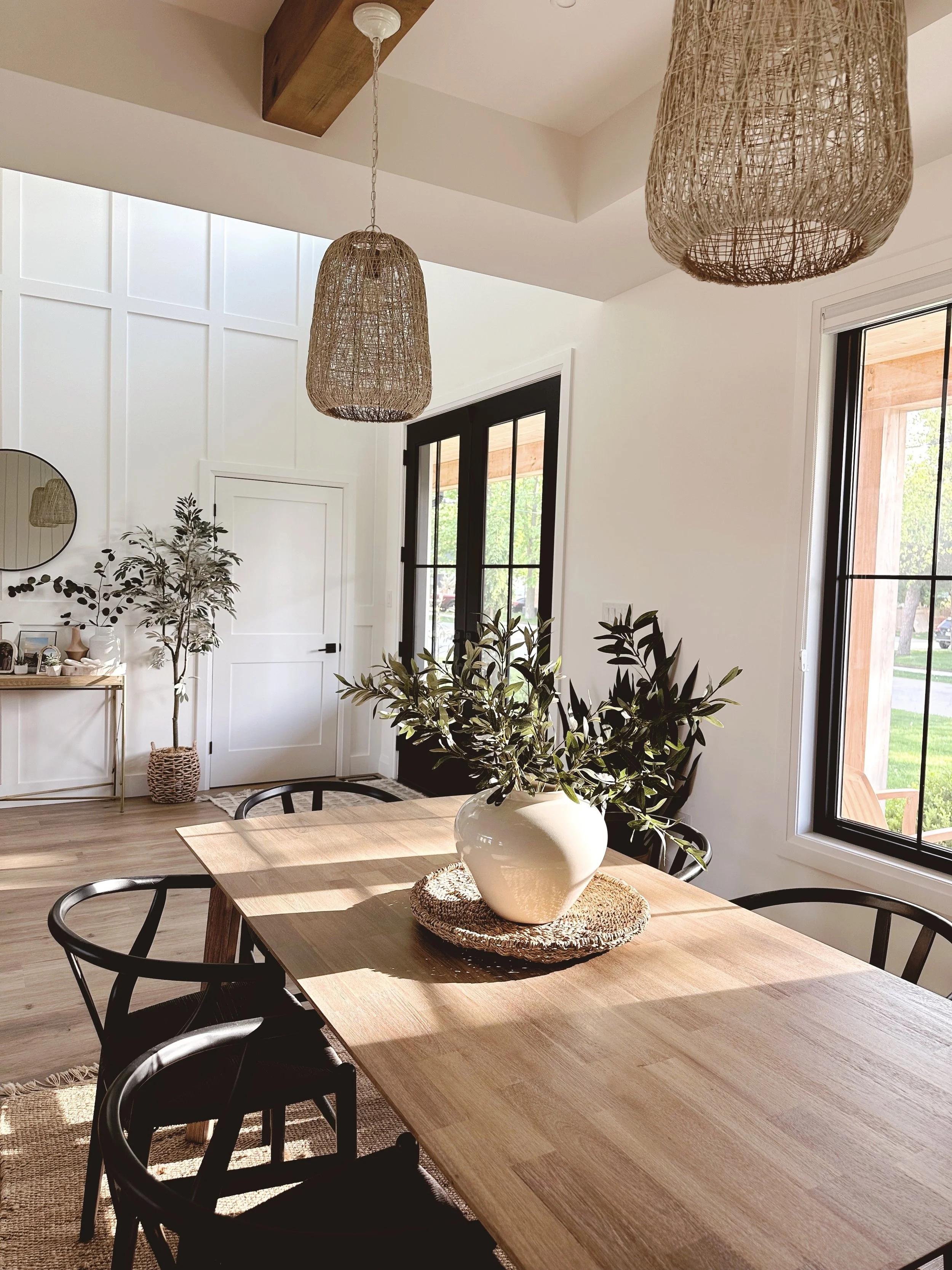 A dining room with a wooden table, black chairs, a white vase with greenery, large windows, and woven pendant lights.