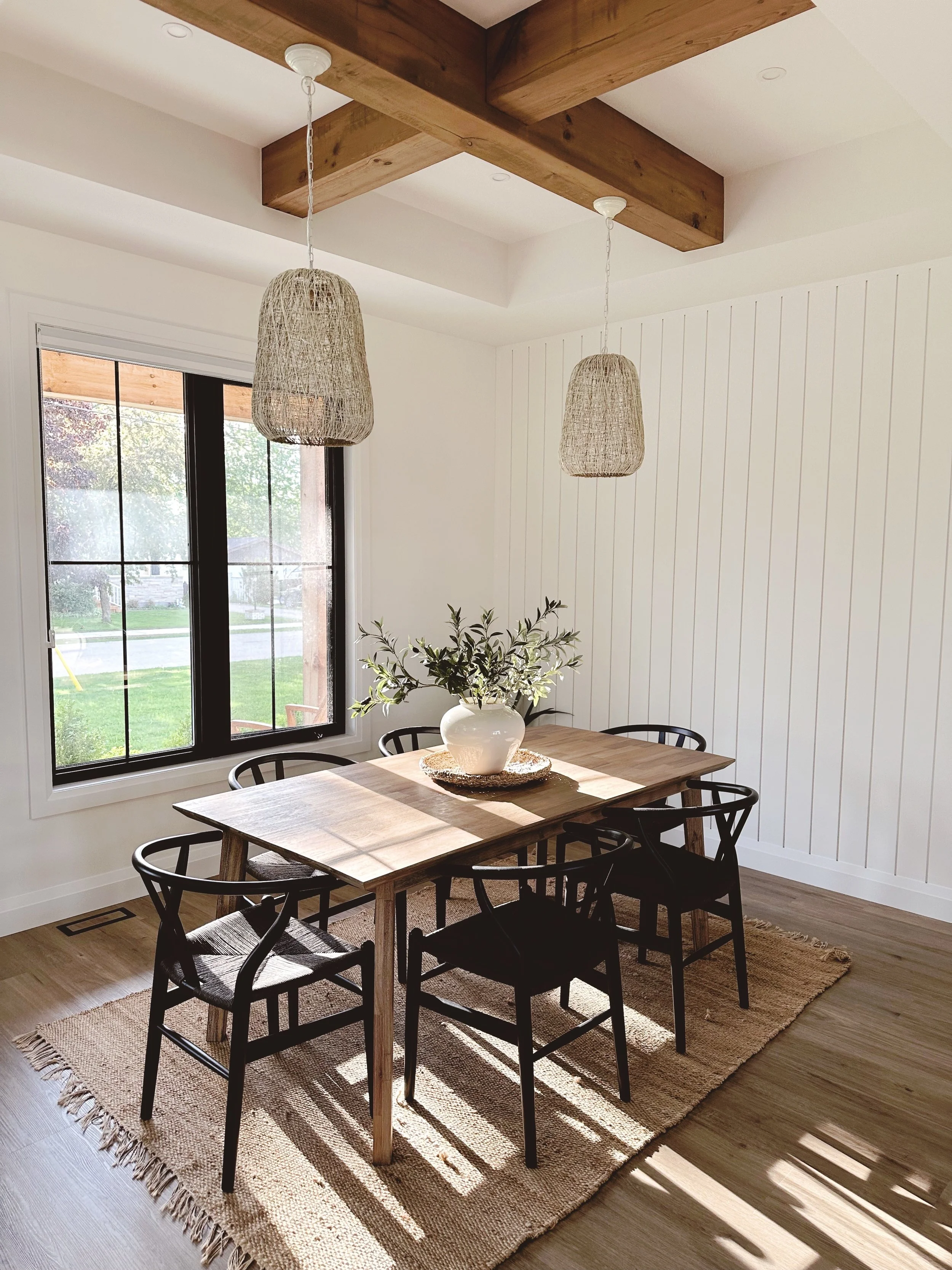 Interior of a dining room with a wooden table, six black chairs, a large window showing a view of a lawn, curtains, hanging wicker pendant lights, a white vase with a leafy plant, and a beige rug on a wooden floor.