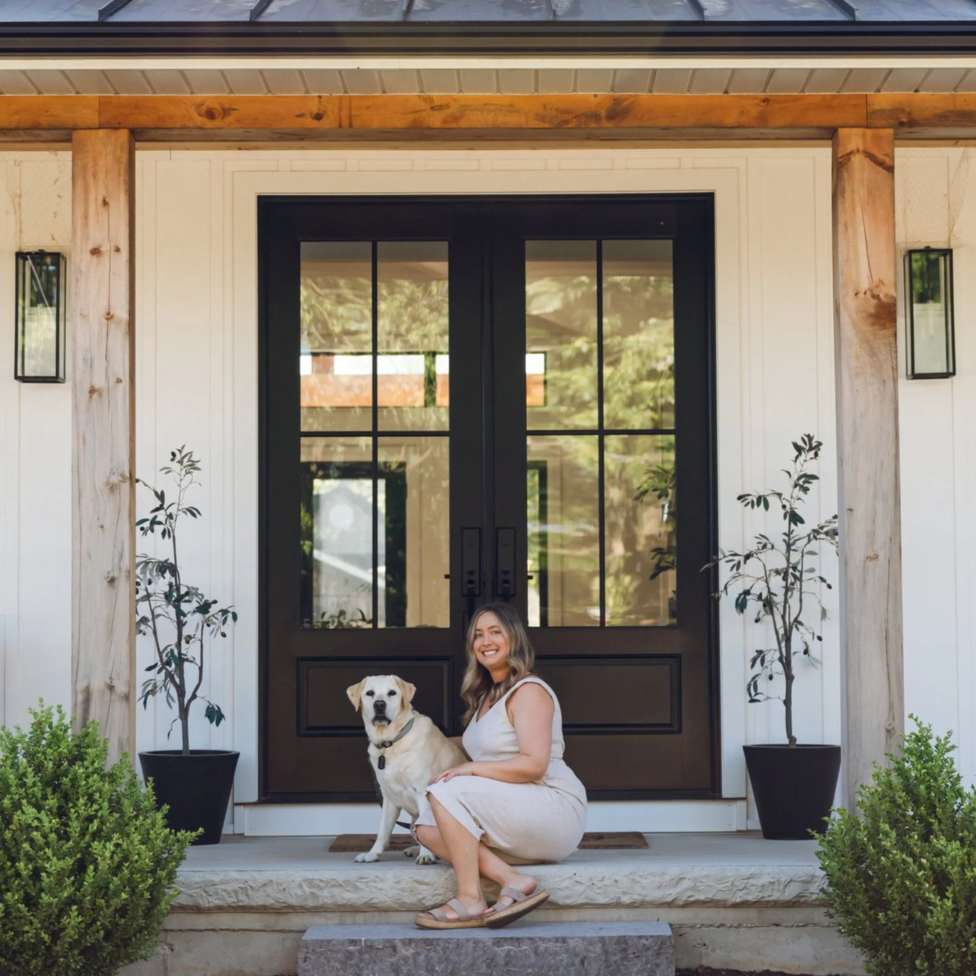 Alyssa and a her dog sitting on the front porch of a house, smiling and looking at the camera. The house has a black door with glass panels, flanked by potted small trees and greenery.