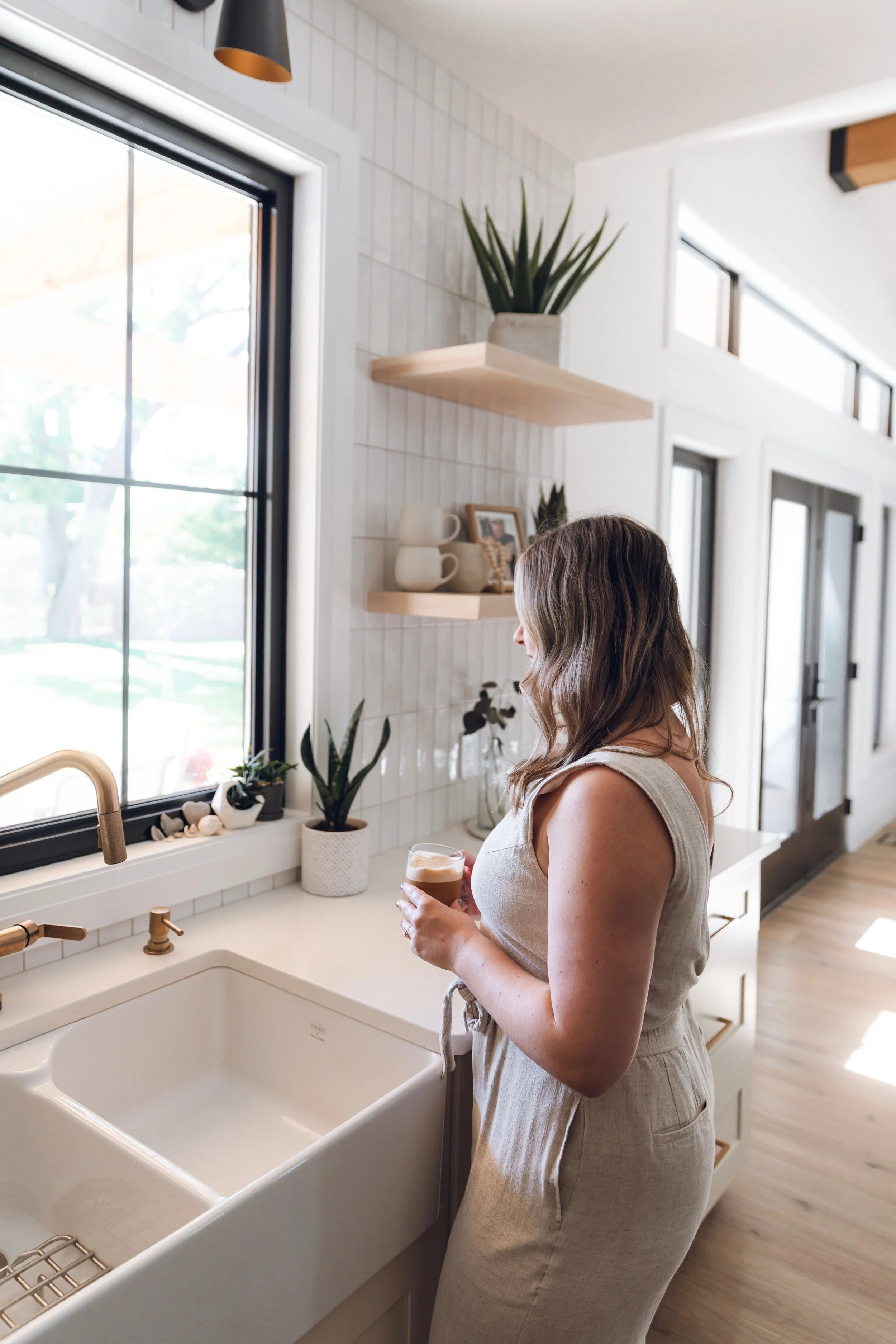Alyssa in a light beige linen dress holding a glass of coffee or tea, standing in a bright kitchen by a farmhouse sink with a gold faucet, near a stovetop, with plants and shelves on the wall behind.