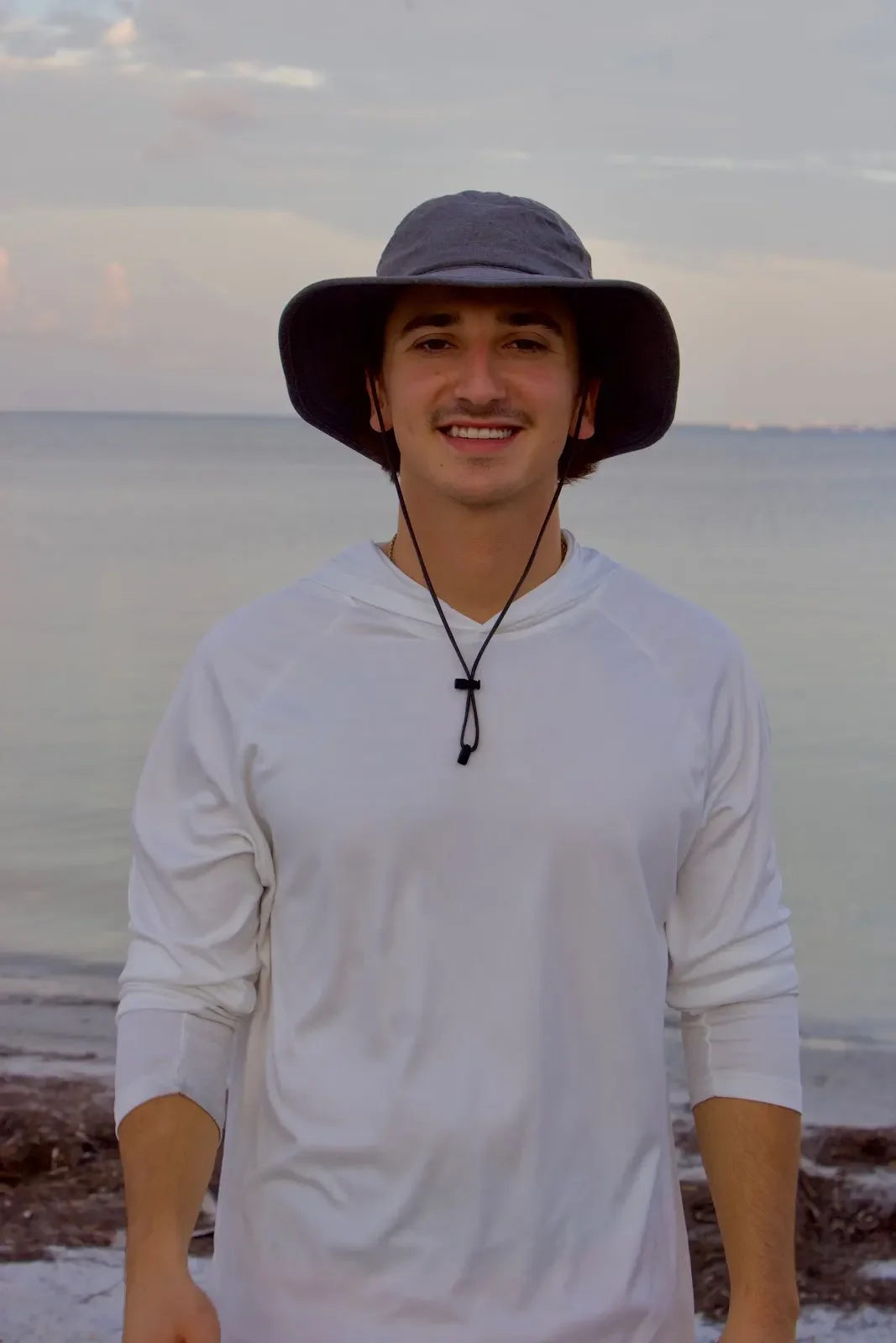 A young man wearing a wide-brimmed hat and a white long-sleeved shirt, smiling on a beach with water and a cloudy sky in the background.