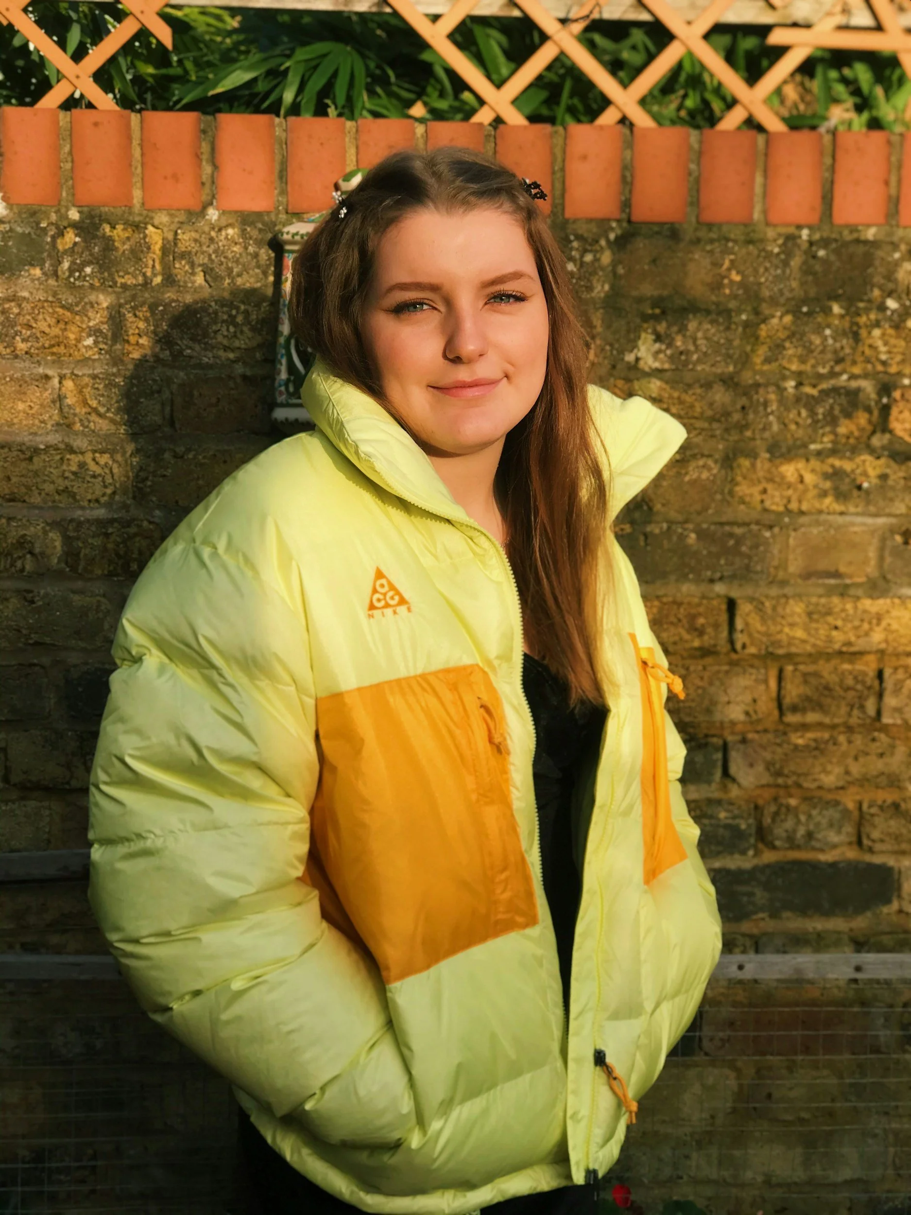 A young woman with long brown hair, blue eyes, and fair skin, smiling slightly, wearing a bright yellow and orange Nike jacket, standing outdoors against a background of brick and greenery.