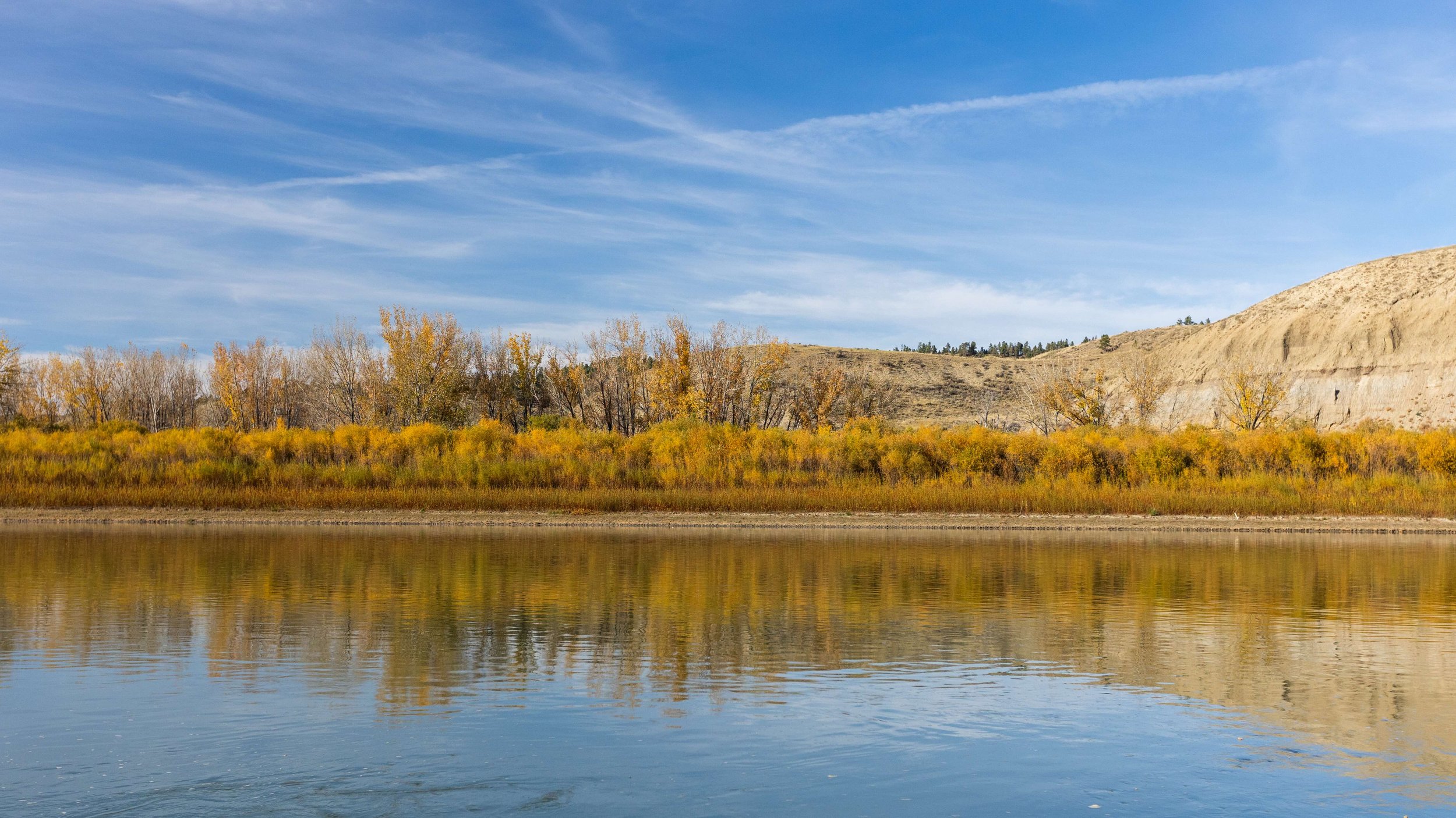 A landscape scene with a calm body of water reflecting the yellow and green trees along the shoreline, a hillside on the right, and a blue sky with wispy clouds above.