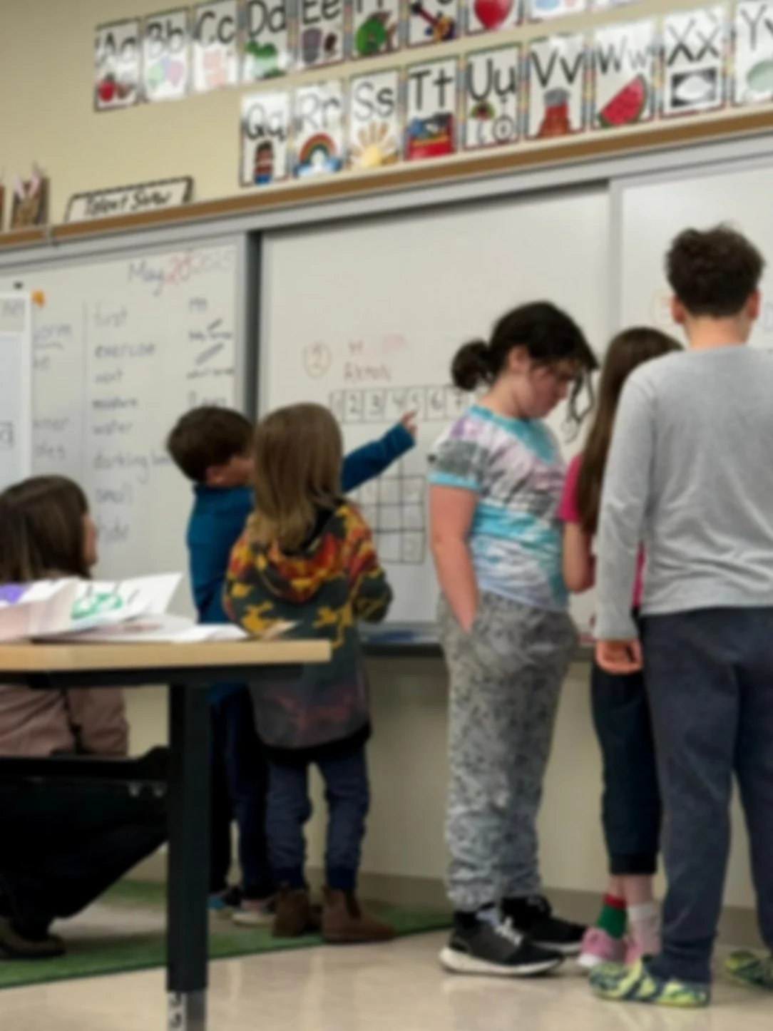 Group of children working together at a whiteboard in a classroom, with colorful educational posters and alphabet charts on the wall.