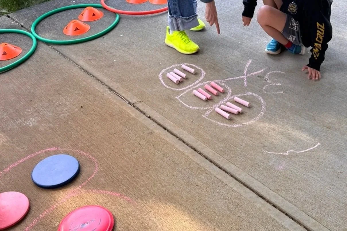 Children playing hopscotch on a concrete sidewalk with chalk, cones, and hoops nearby.