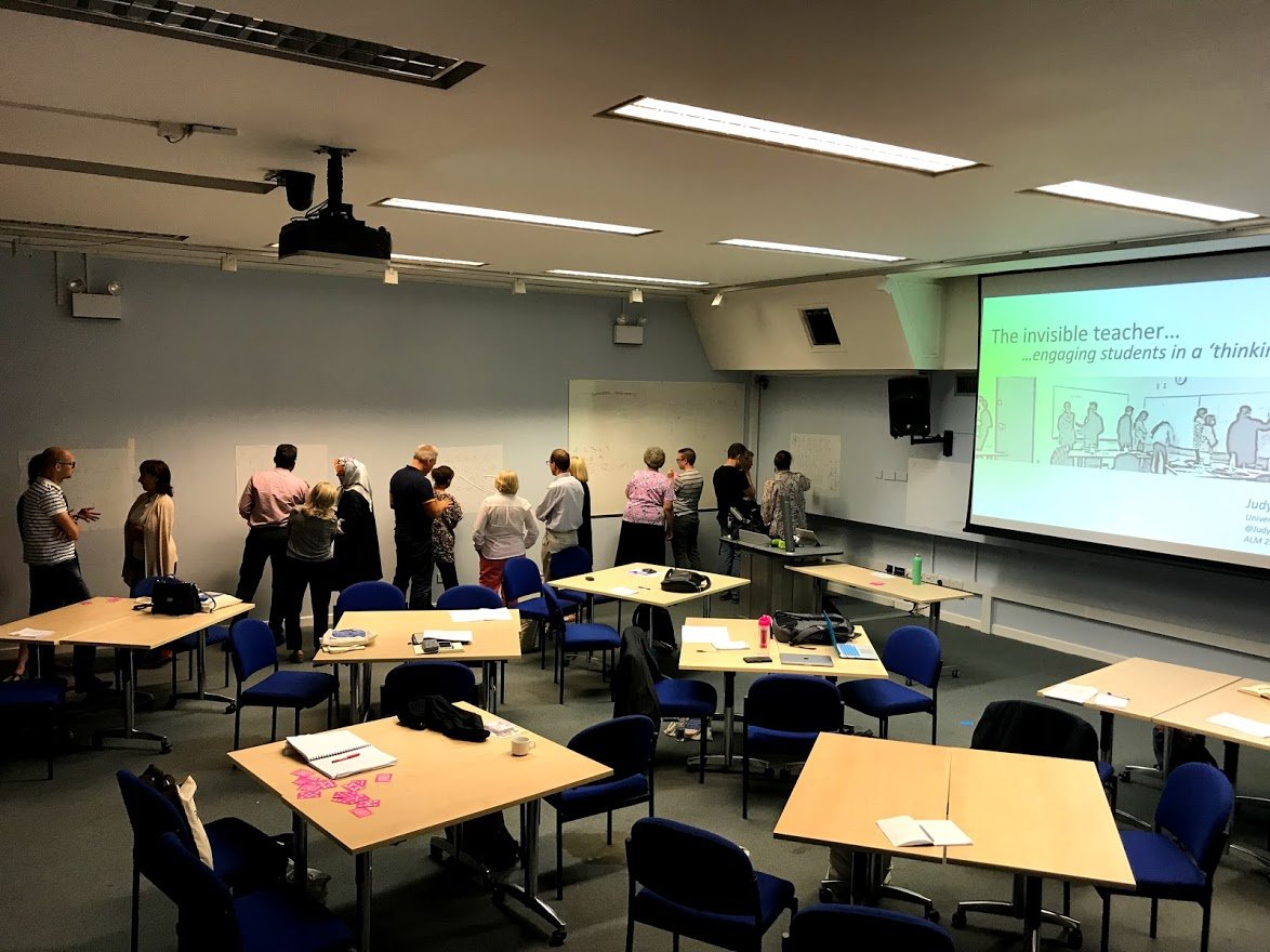 A classroom or conference room with tables and chairs, some people standing near whiteboards along the back wall, and a large projection screen displaying a presentation slide.