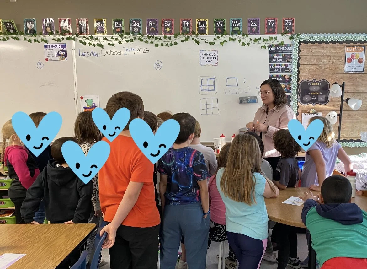A classroom with young students gathered around a teacher at a whiteboard, which displays handwritten notes and diagrams. The classroom has colorful alphabet banners along the top, and various educational posters on the walls.