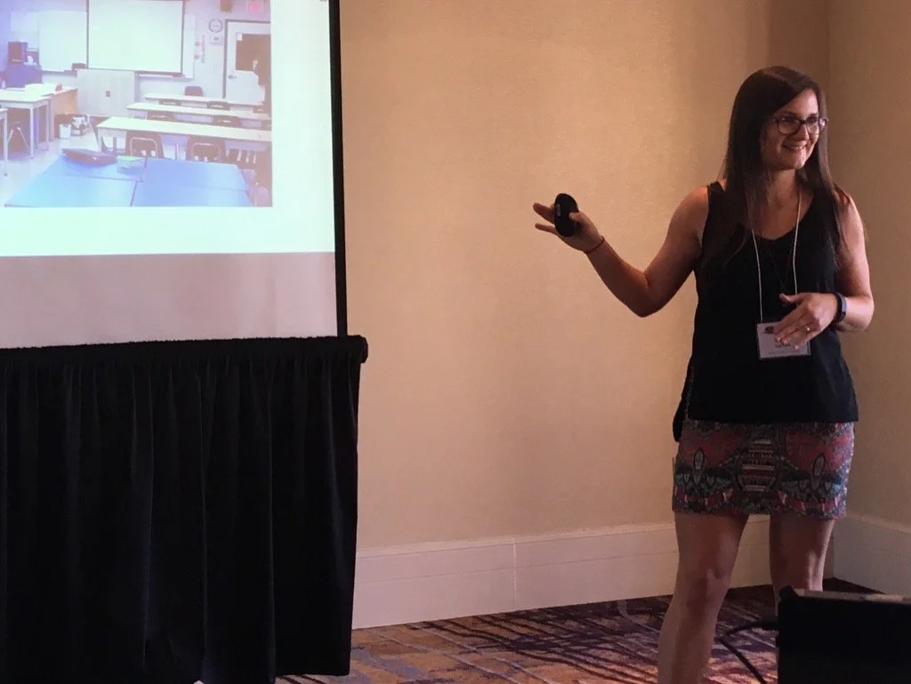 Woman giving a presentation in a conference room with a projected slide of an empty classroom.