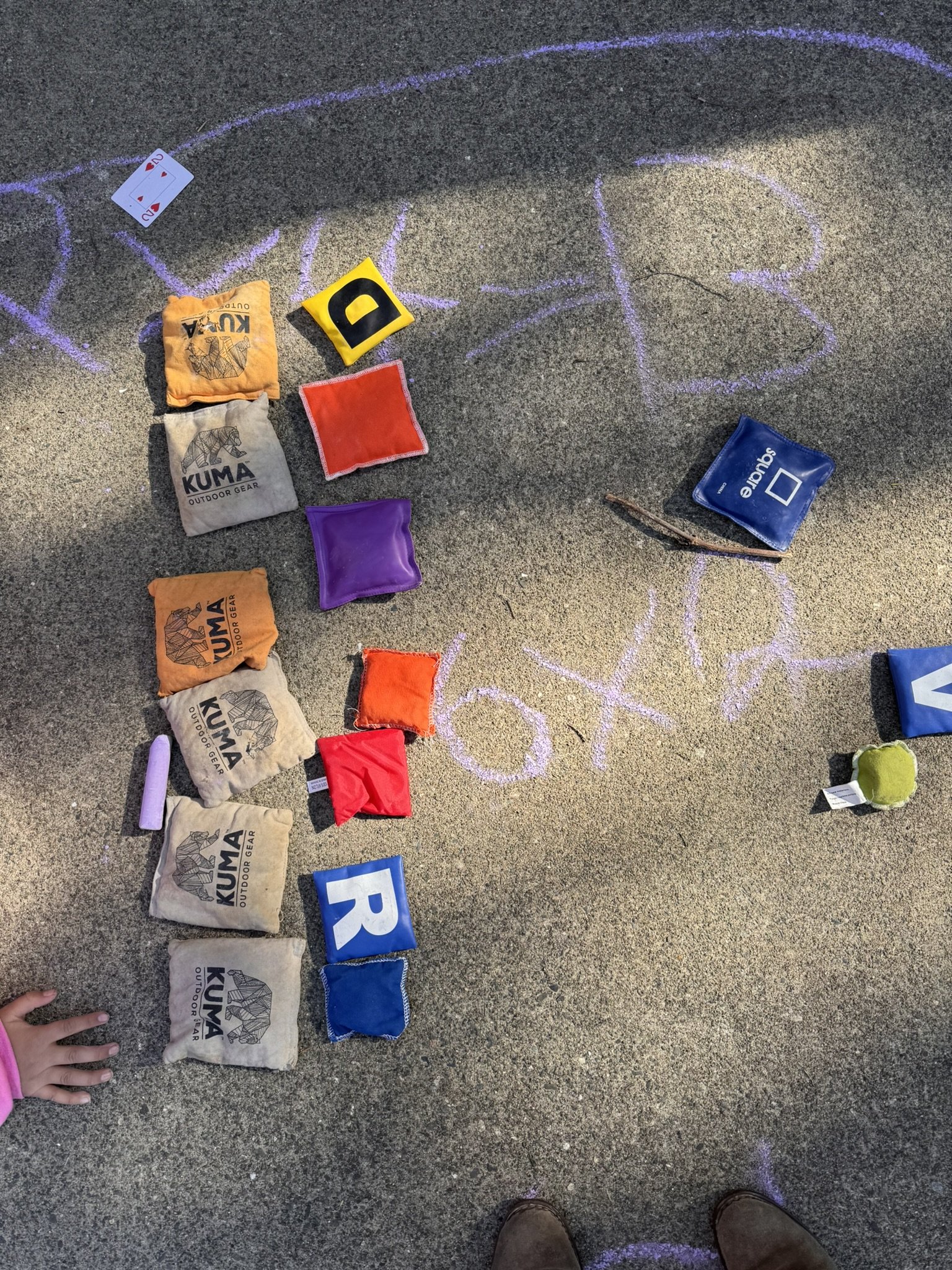 Colorful bags and cloths with letters and chalk drawings on pavement, with a child's hand and shoes visible at the bottom of the image.