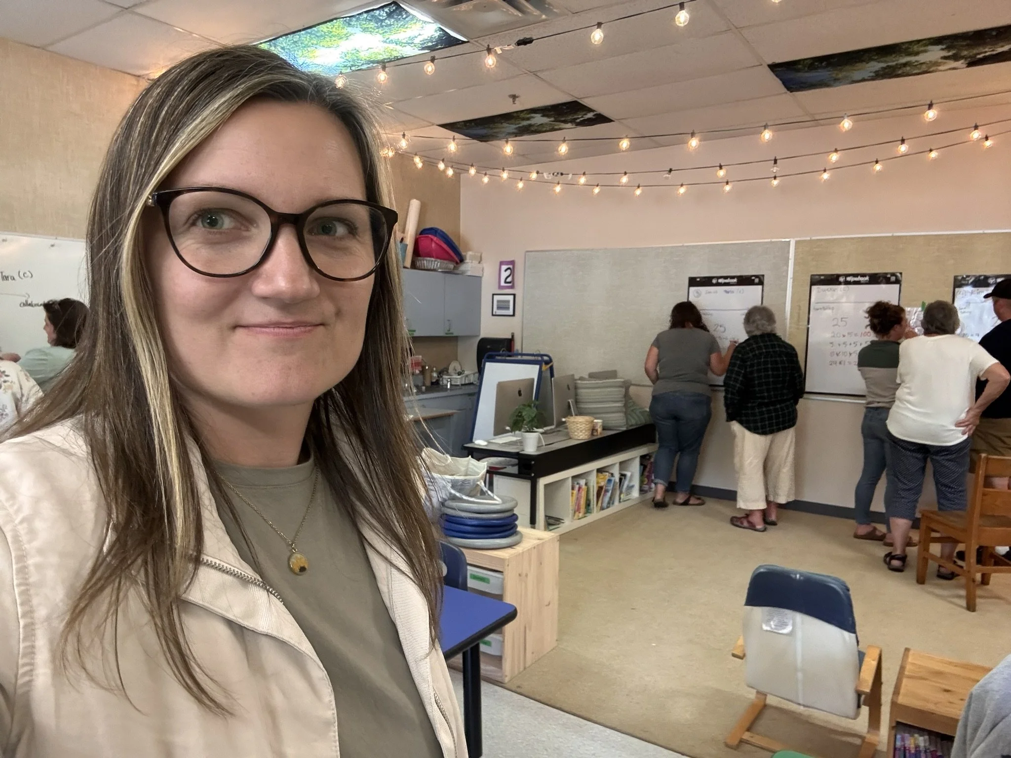 A woman with glasses and long hair in the foreground of a classroom setting, with several people and whiteboards with writing in the background, and ceiling tiles decorated with outdoor scenery and string lights overhead.
