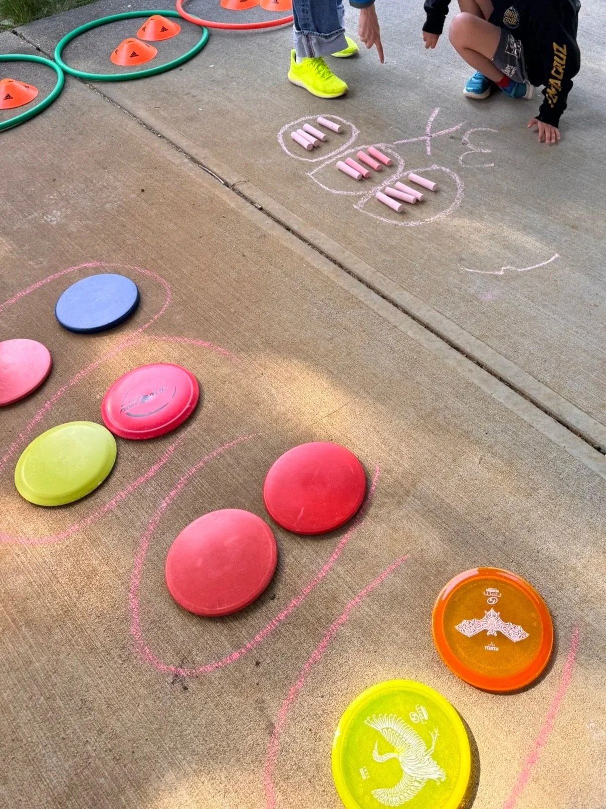 Children playing disc golf outdoors, with colorful discs on the ground, cones and hula hoops for training, and chalk markings on the concrete sidewalk.