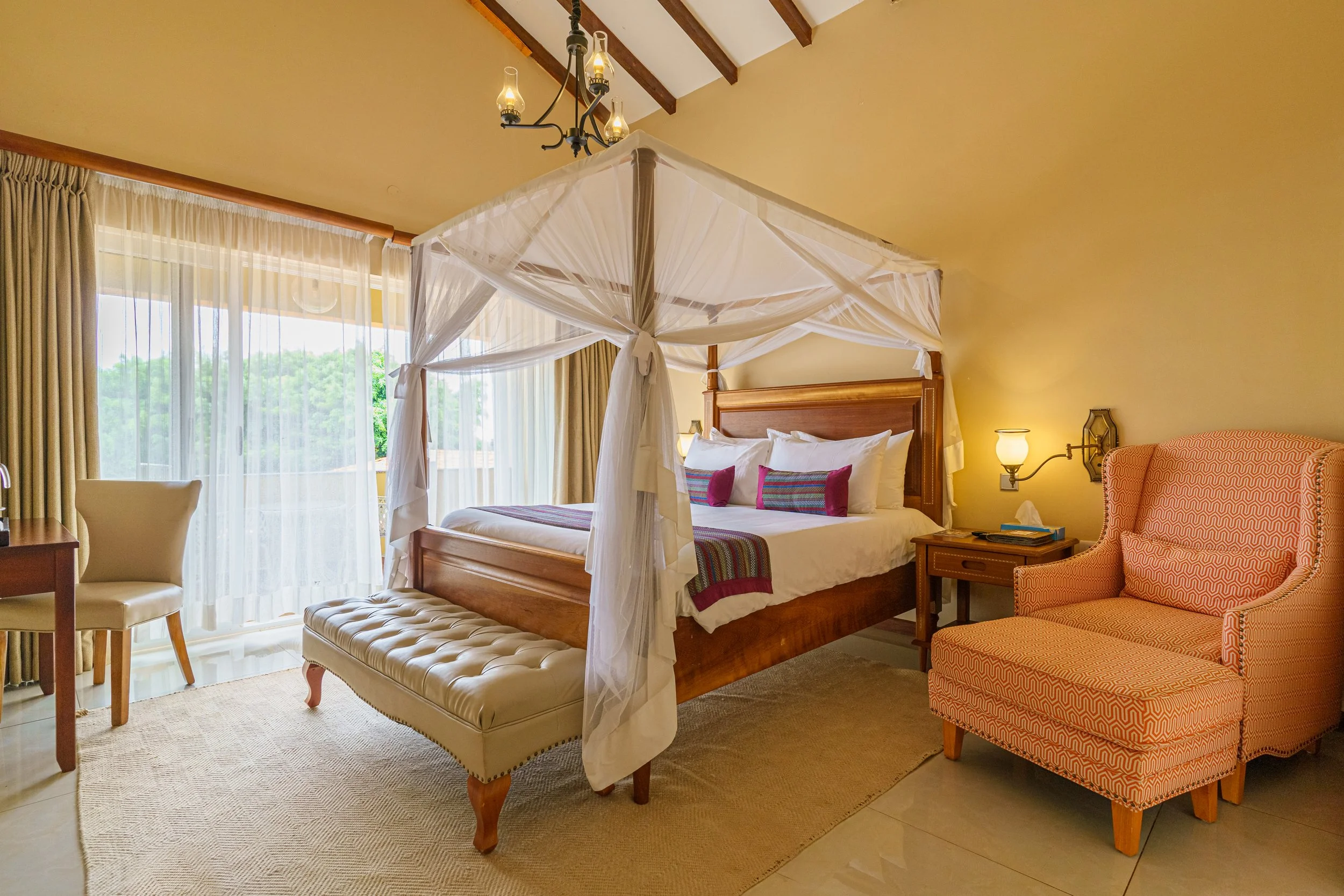 Room with a four-poster bed with white drapes, wooden headboard, colorful pillows, and a matching bench at the foot. To the right, an orange patterned armchair and small side table with a lamp. To the left, a cream-colored chair and a view of large windows with sheer curtains showing an outdoor balcony and green scenery.