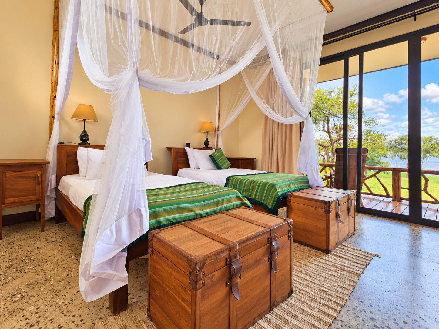 A bedroom with two beds featuring green striped bedding, wooden furniture, and a canopy mosquito net. There's a sliding glass door leading to a balcony with a scenic view of trees and a lake or river.