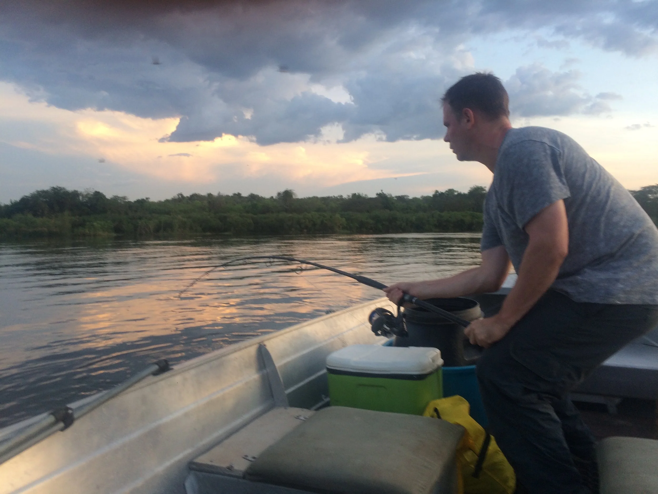 A man fishing on a boat during sunset, with dark clouds and a water body and greenery in the background.