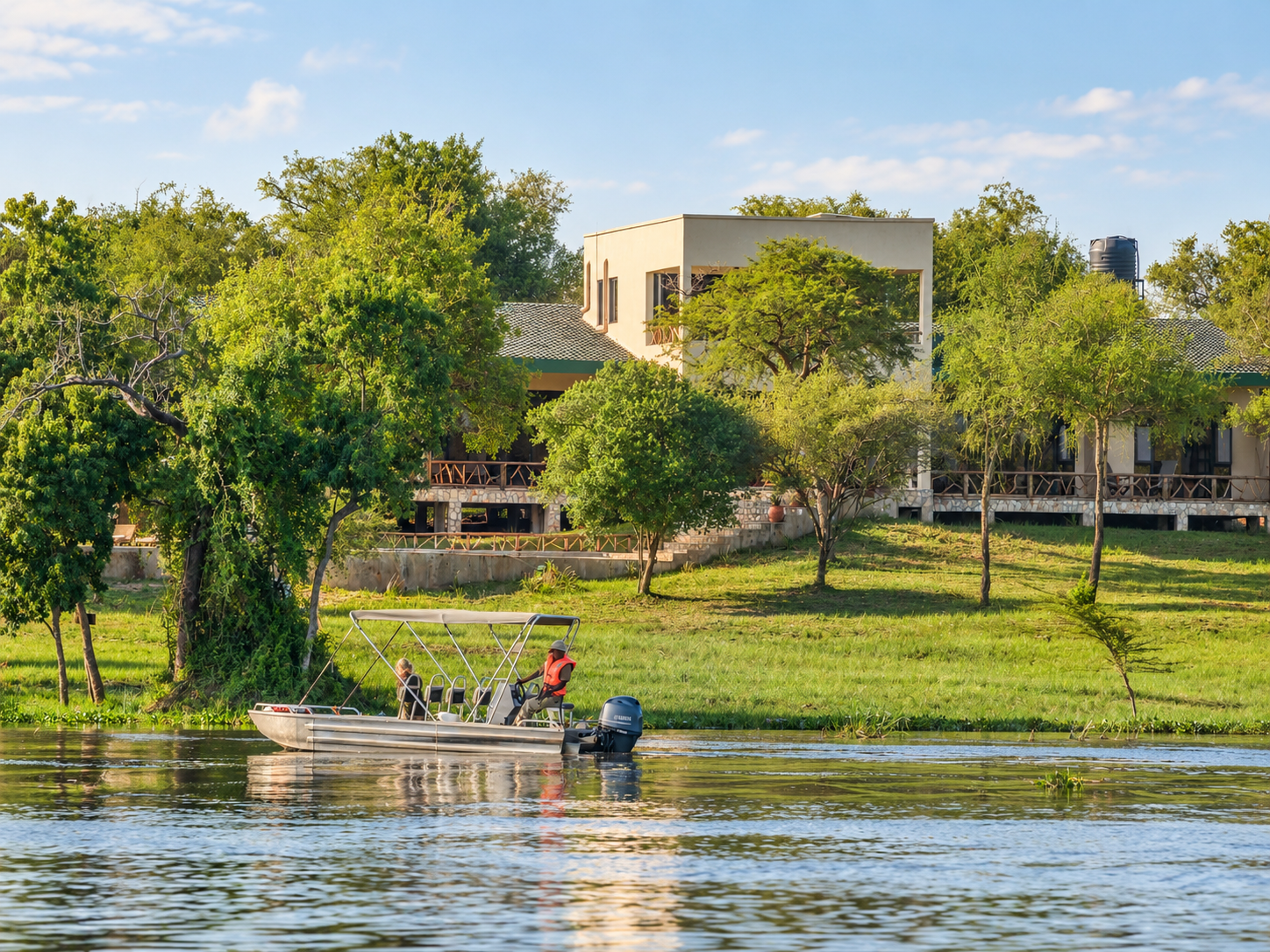 A boat with two people on a river next to a grassy bank with trees and a house in the background under a blue sky.