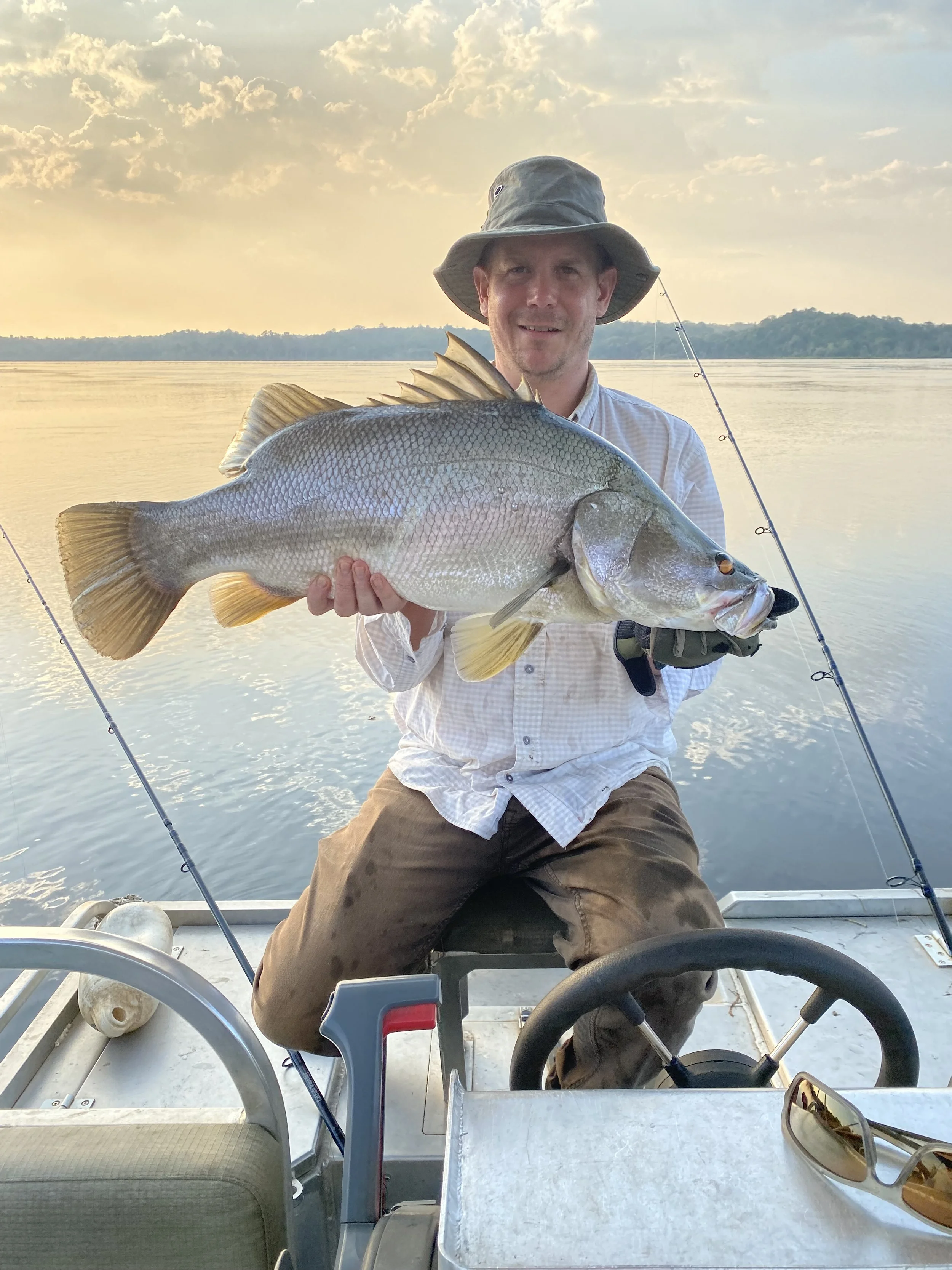 A man sitting on a boat holding a large fish in his hands, wearing a gray hat and a beige shirt, with fishing poles in the background on a body of water at sunset.