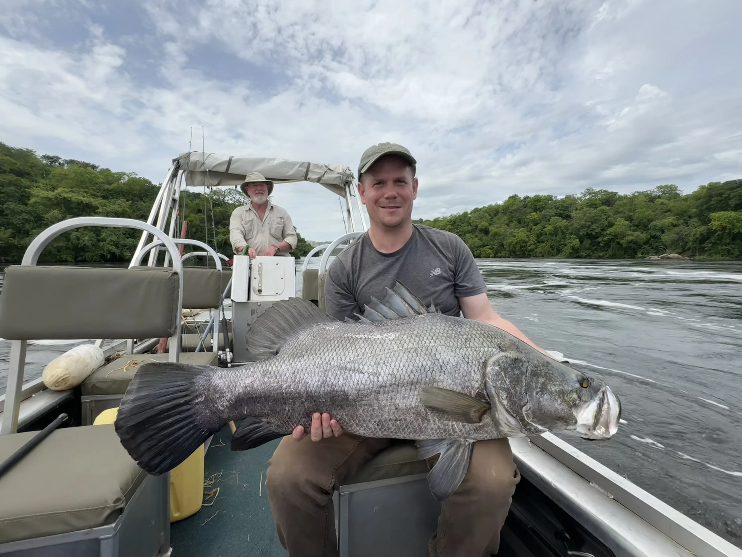 A man holding a large fish on a boat with two men and green trees in the background under a cloudy sky.