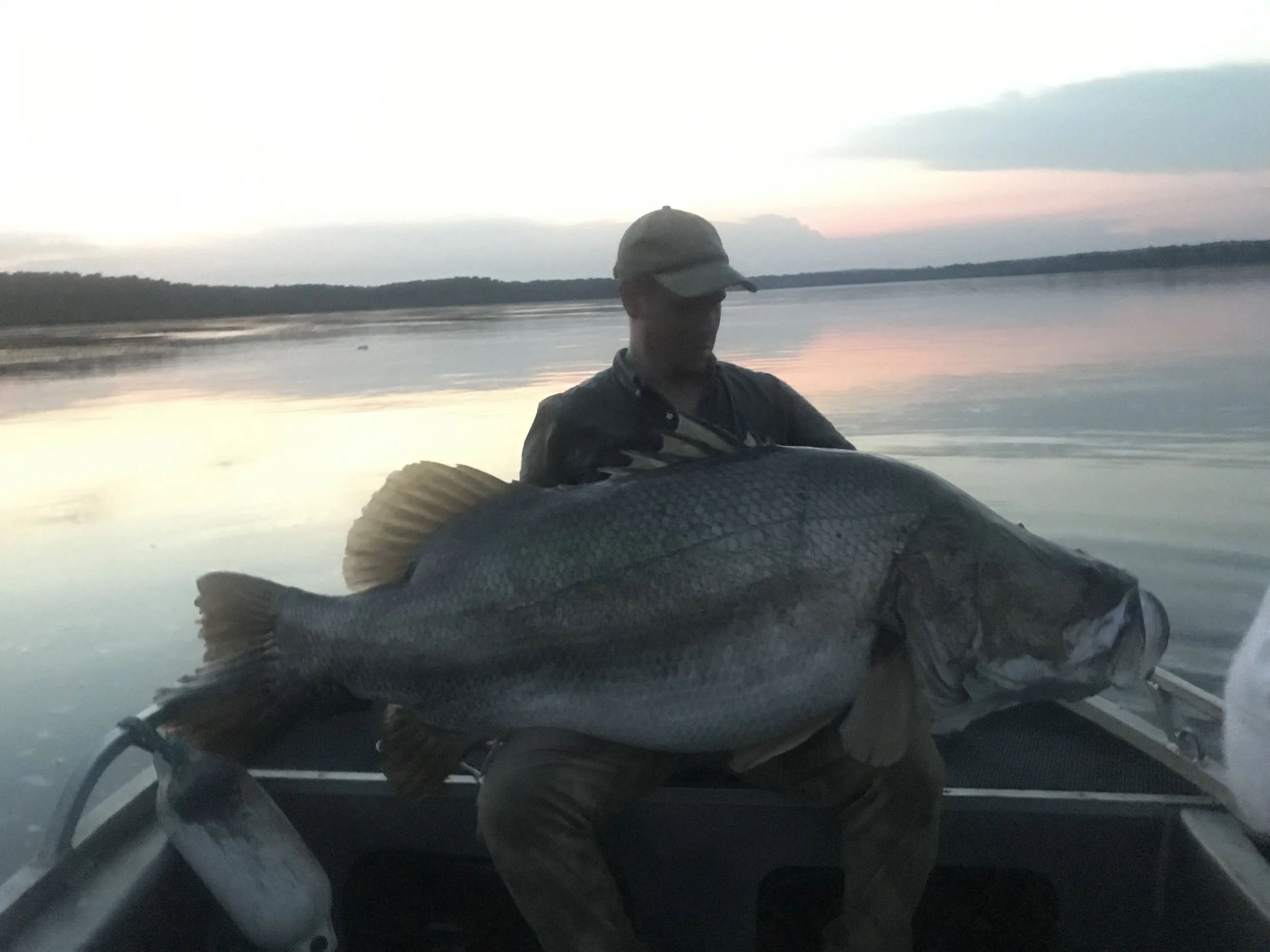 A person with a hat and dark clothing in a boat, holding a large fish, with a body of water and a sunset or sunrise in the background.