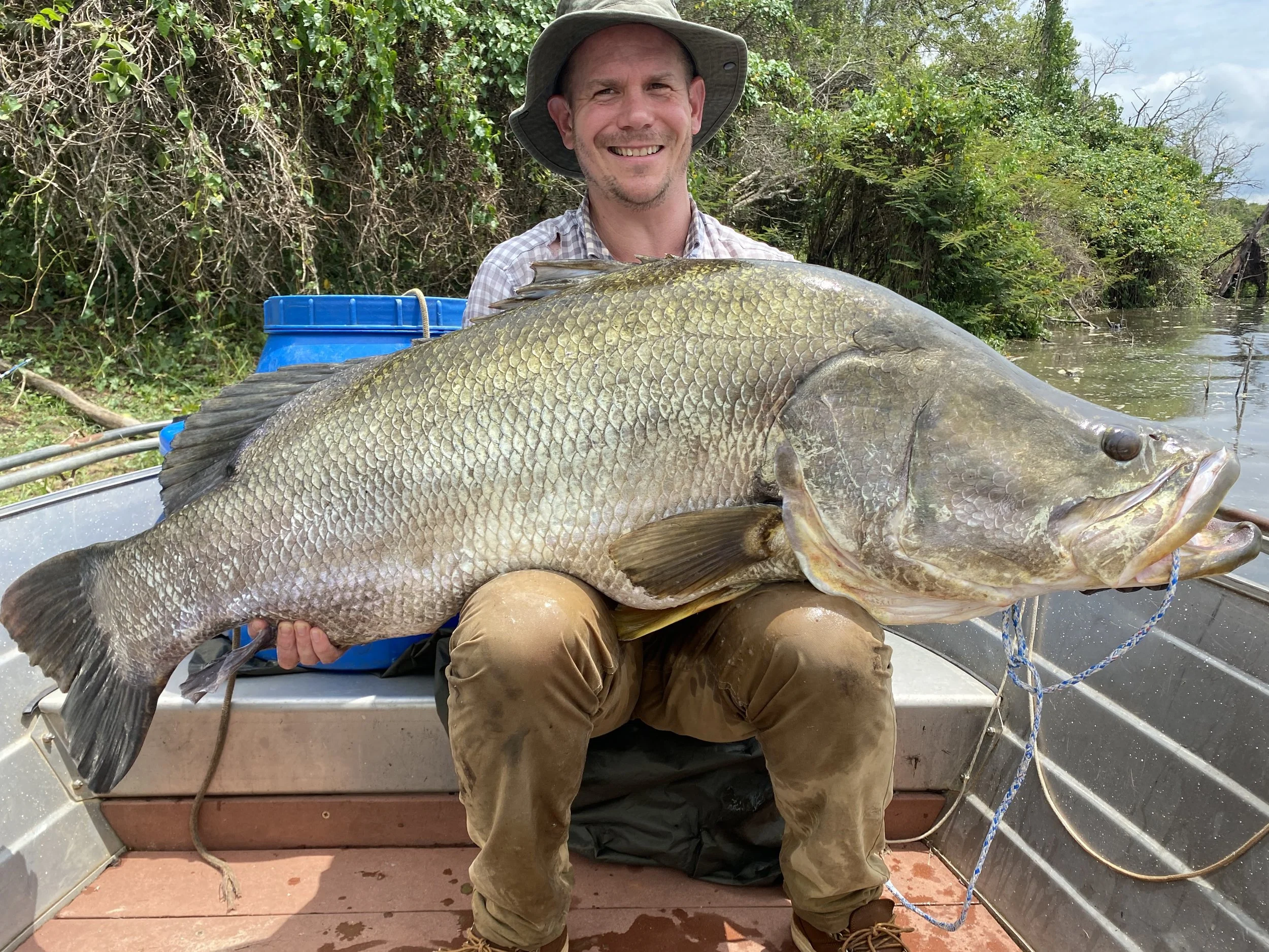 A man sitting in a boat holding a large fish, with a green, wooded shoreline behind him.