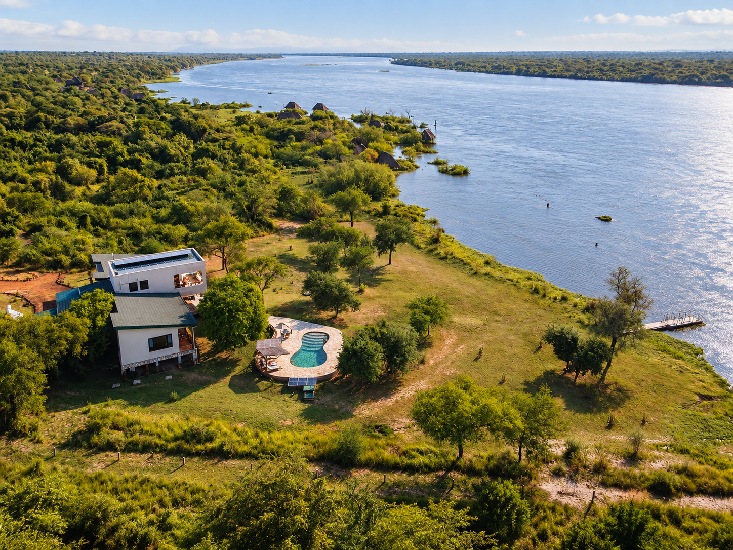 Aerial view of a lakeside property with modern house, swimming pool, surrounded by greenery and trees, with small dock on the water and lush landscape extending to the horizon.