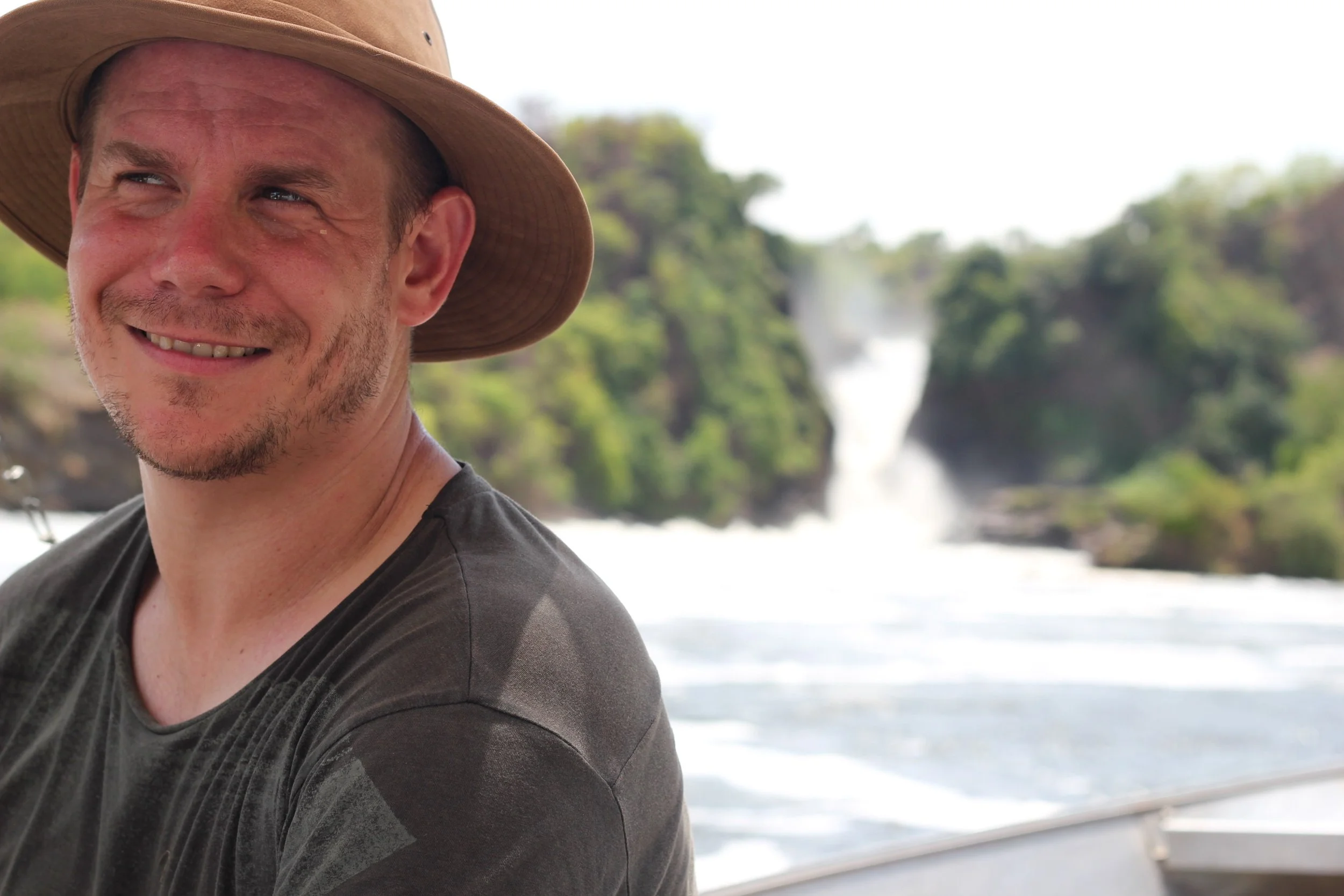 A smiling man wearing a wide-brimmed hat and a dark t-shirt outdoors near a waterfall and lush green trees.