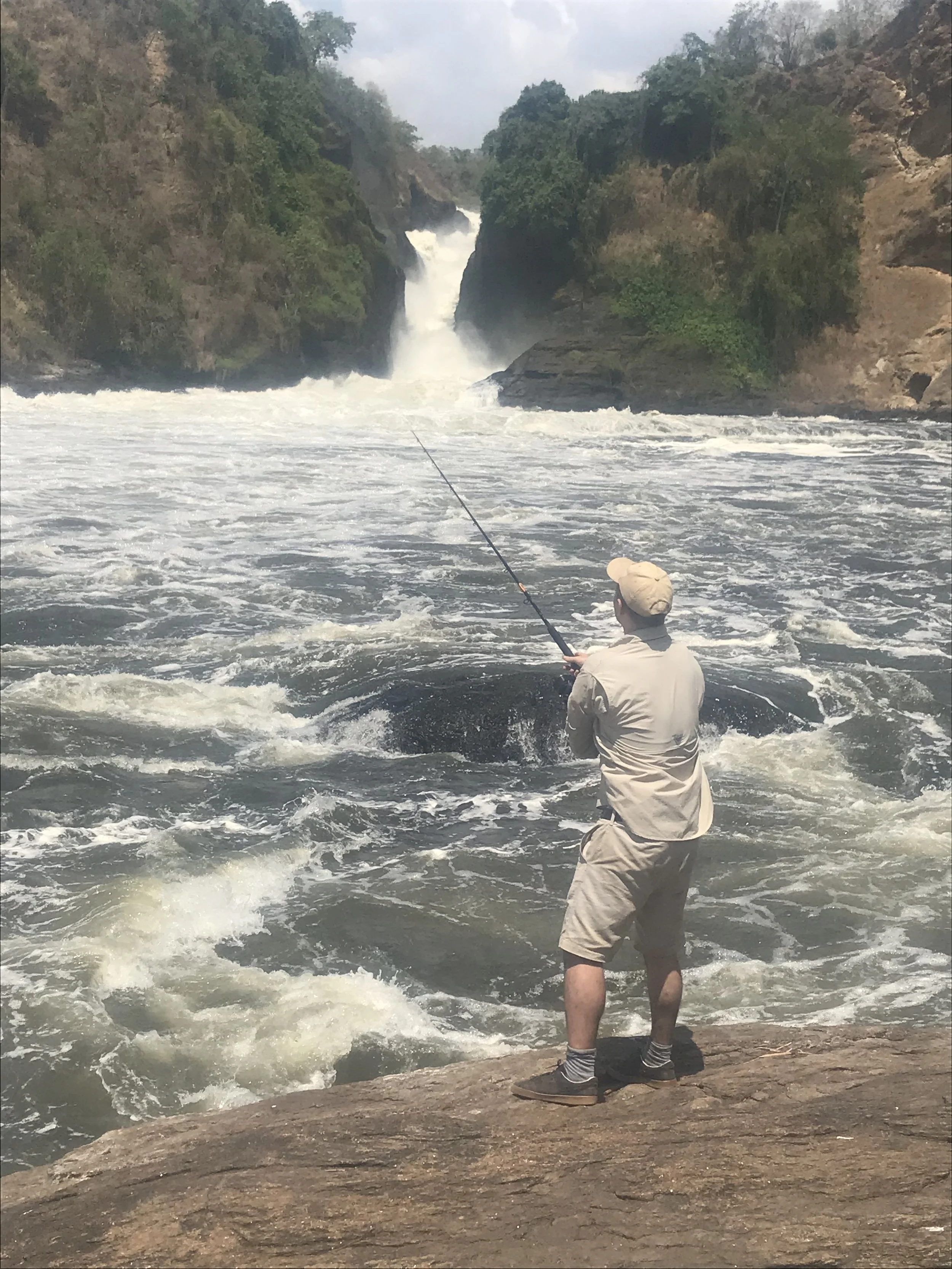 Person fishing in a river near a waterfall surrounded by trees and rocky cliffs.