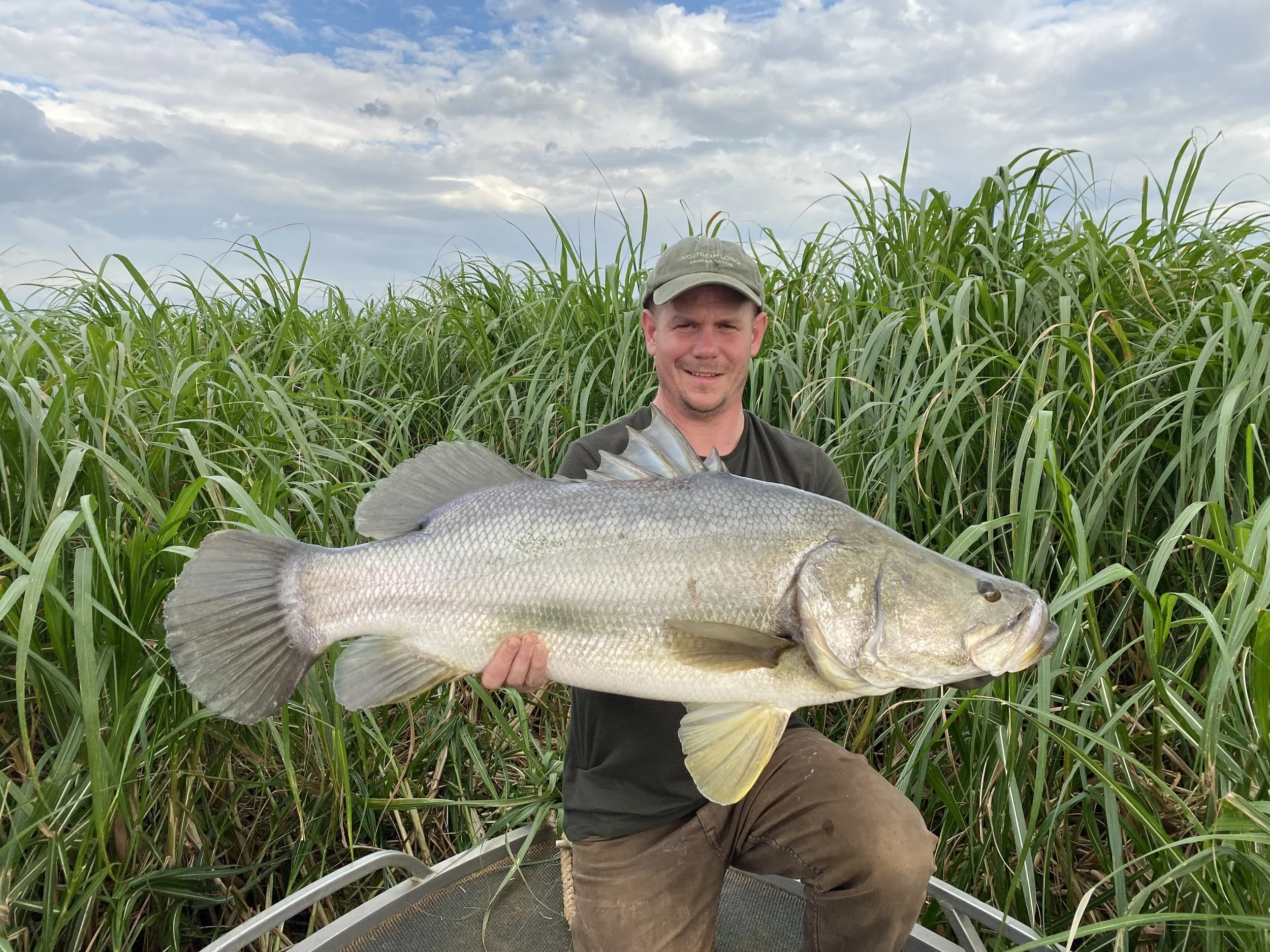 A man wearing a baseball cap and dark clothing holding a large fish in front of tall green grass under a partly cloudy sky.