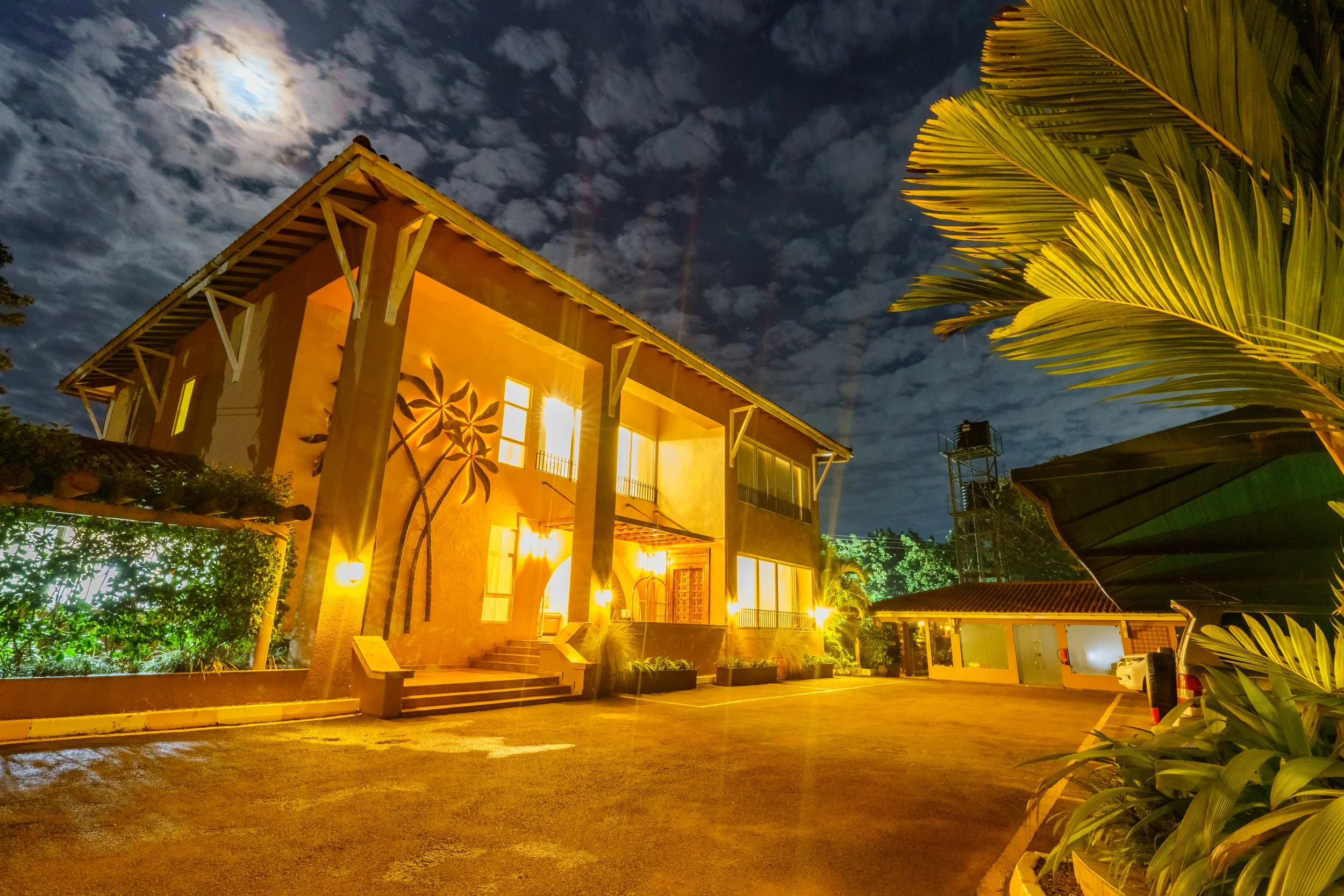 Nighttime view of a house with warm yellow lighting, the moon partially covered by clouds, a large palm frond on the right, and a parking lot with cars in the foreground.