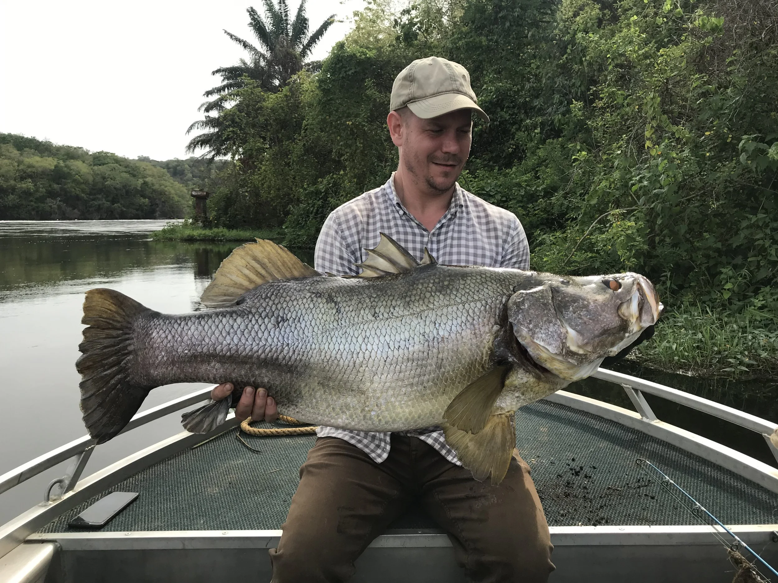 Angler holding a giant Nile perch aboard a fishing boat on Uganda's River Nile, with green forest along the riverbank.